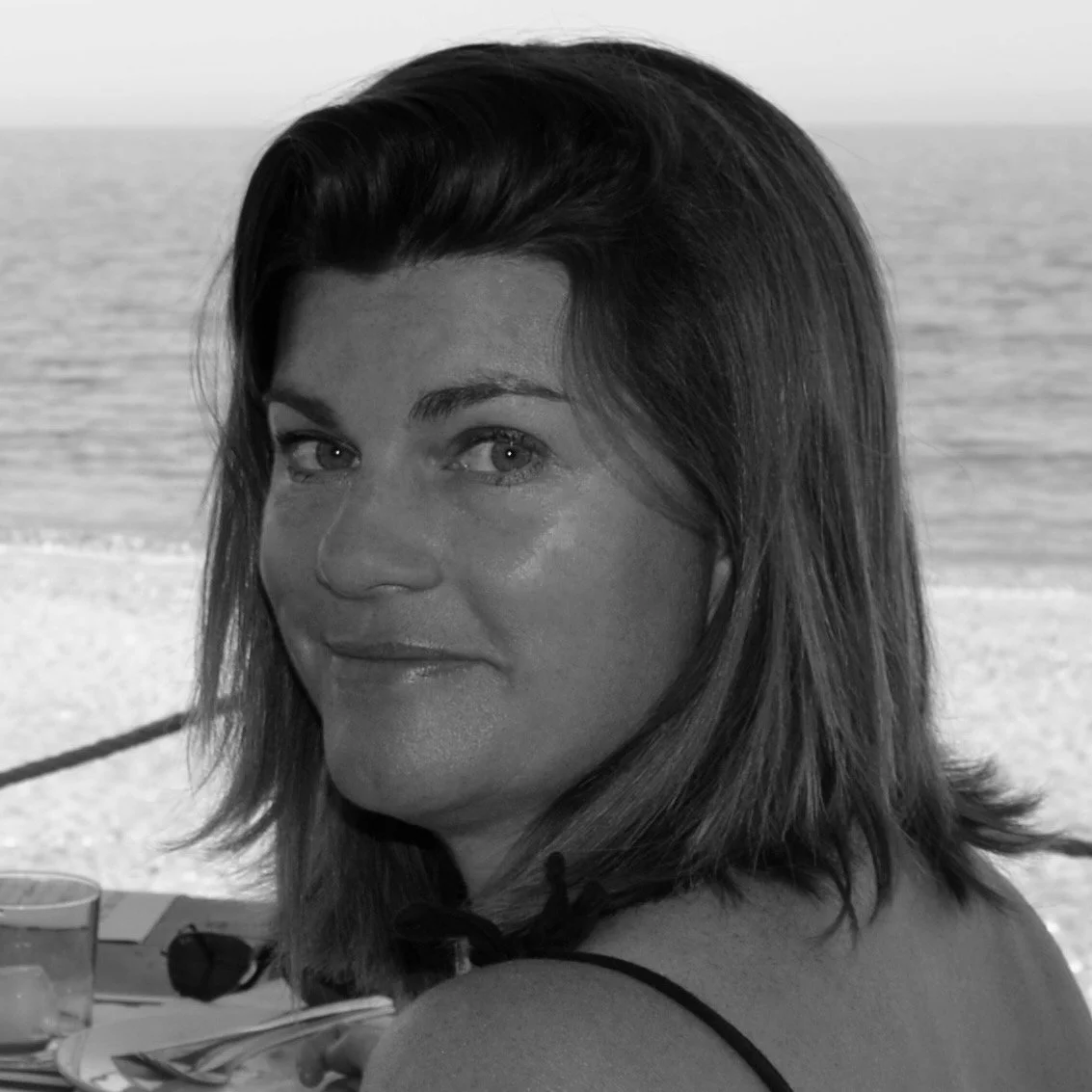 Black-and-white photo of a woman with shoulder-length hair, sitting on a beach, with the ocean in the background. Soft Tissue Therapist. musculoskeletal specialist.