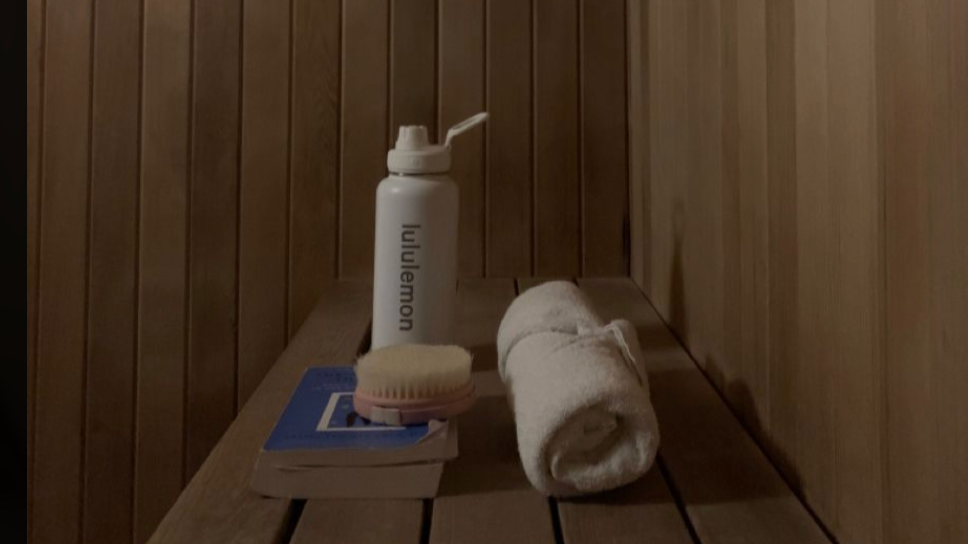 A wooden table with a white water bottle, a rolled white towel, a closed book, and a white brush. Sussex Infrared Private Sauna.