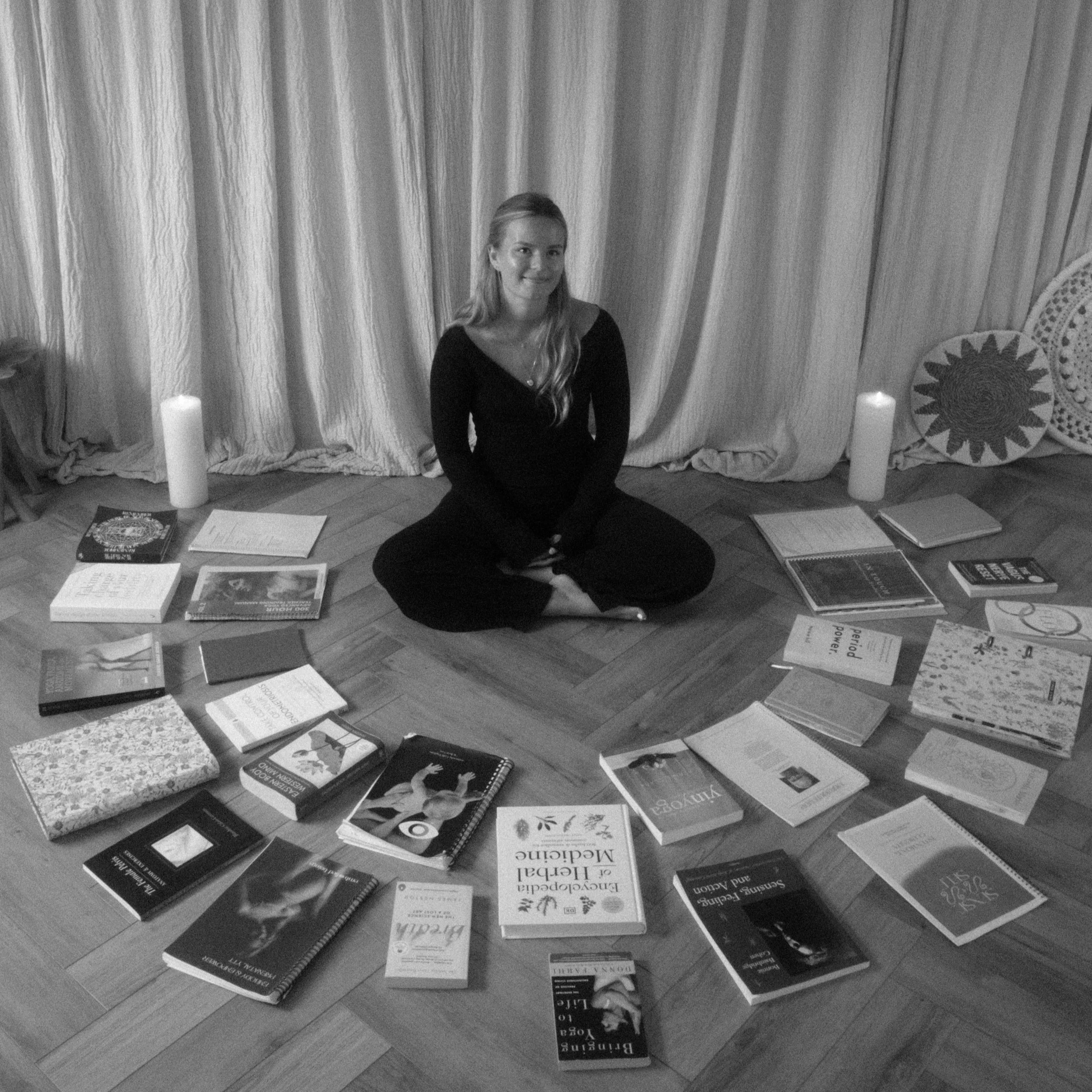Black and white photo of a woman with long hair, sitting on the floor surrounded by books and papers, in a room with curtains and candles. Yoga. Somatics. Nervous System Regulation. Embodiment. Ayurvedic Medicine. Relaxing Massage.