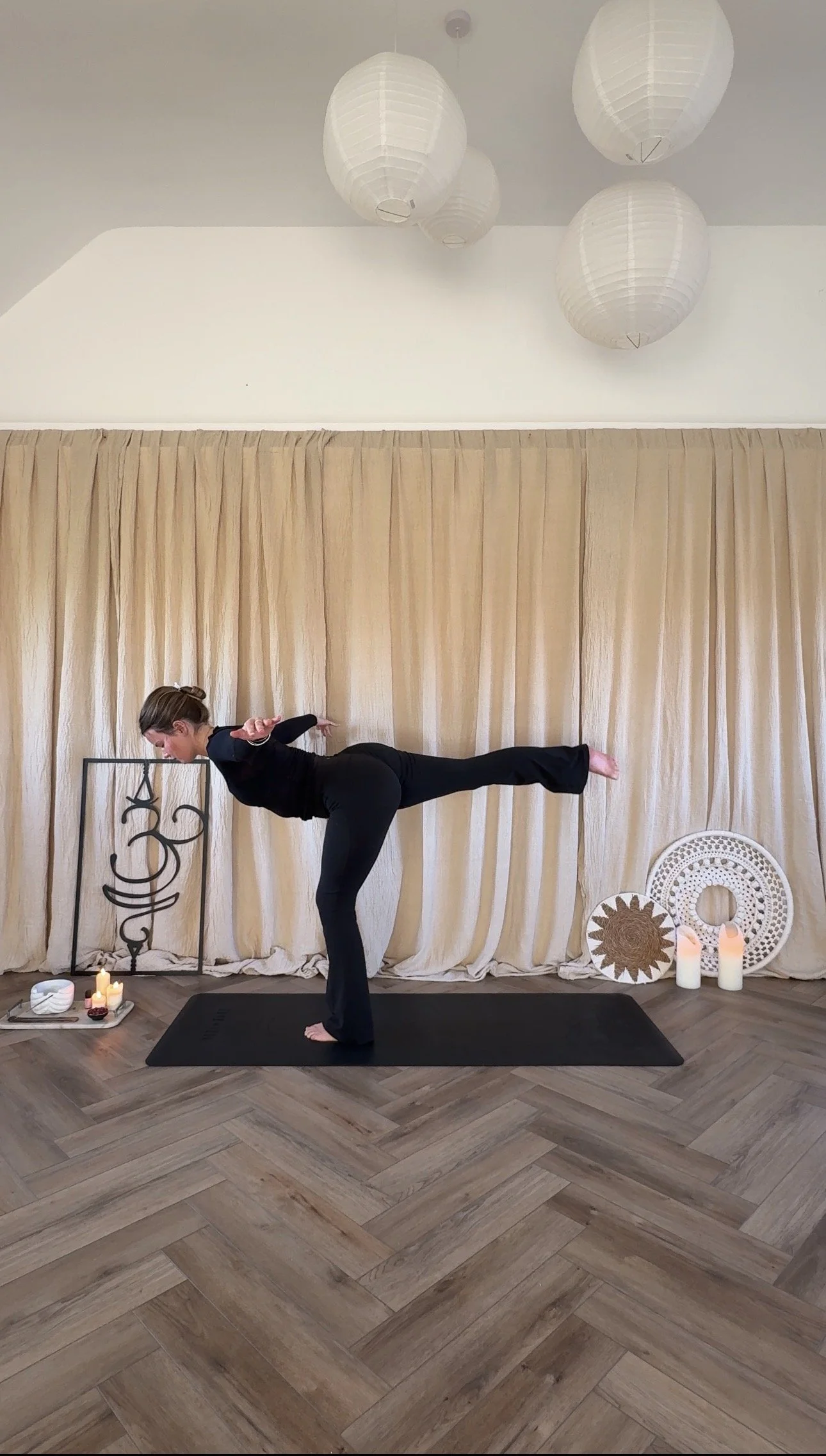 A woman practicing yoga in a indoor studio with beige curtains as background, candles, and decorative items on the floor. She is standing on a black yoga mat, balancing on one leg with the other leg extended behind her, and arms outstretched.