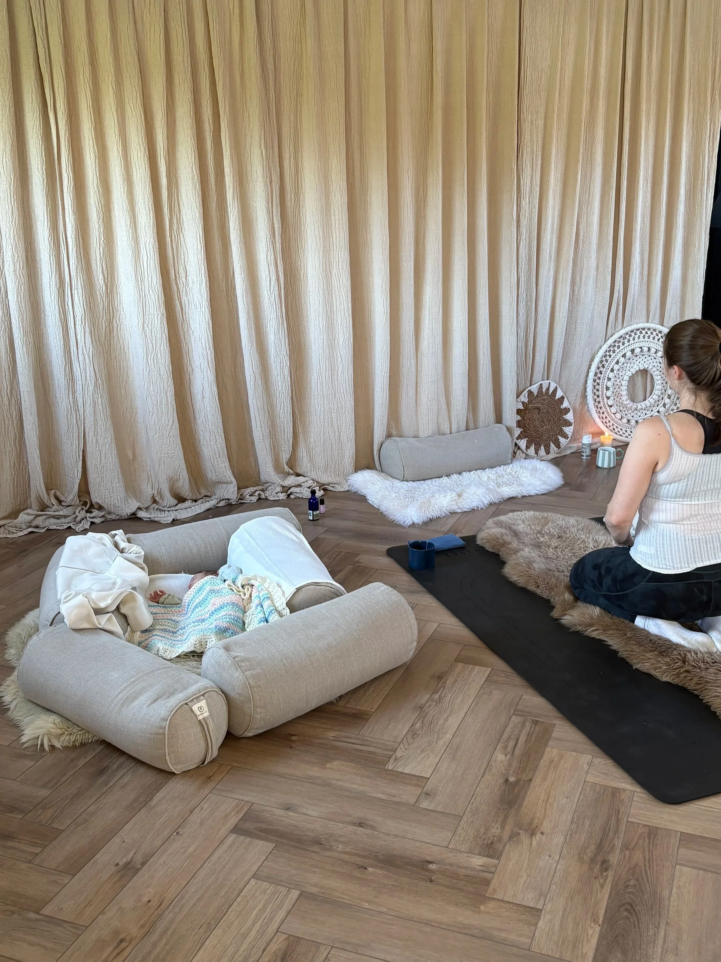 A woman practicing yoga on a black mat with a brown furry blanket, surrounded by decorative cushions, candles, and a baby sleeping in a beige cushioned bed on a wooden floor with beige curtains in the background. Postnatal Yoga