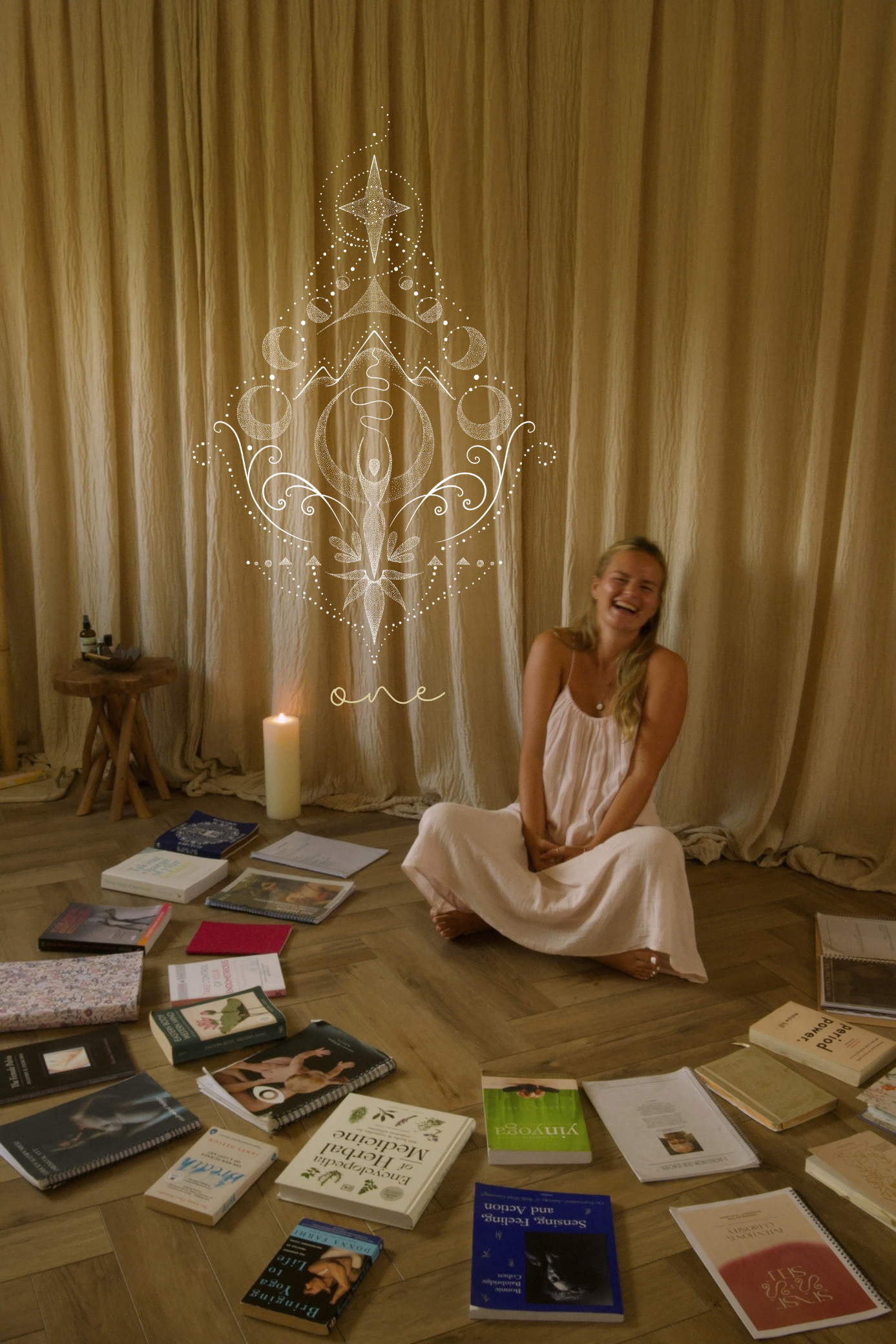 A woman in a white dress sitting cross-legged on the floor, smiling and laughing. Books and magazines are spread around her on the wooden floor. Behind her, a beige curtain covers the wall, with a decorative white graphic overlay resembling a symmetrical design with floral and celestial motifs, and the word 'one' written near the bottom.