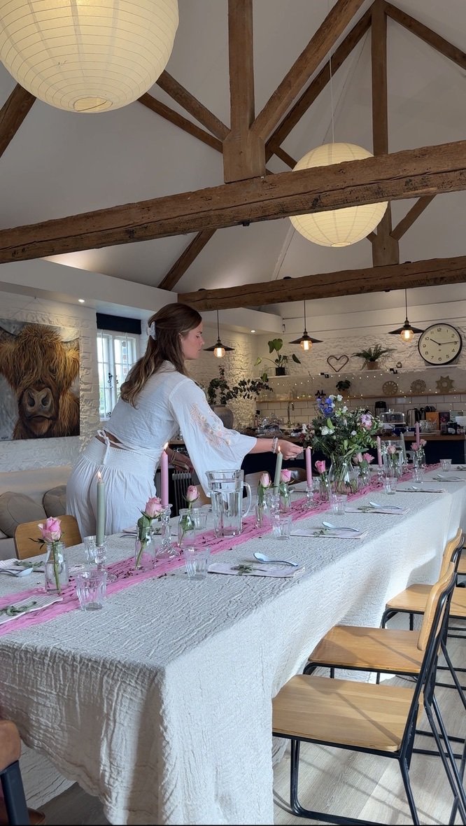 A woman setting a dinner table decorated with pink and white flowers, candles, and glassware inside a rustic room with wooden beams and hanging lamps. Yoga and Brunch yoga retreat at The Boxgrove Clinic in Sussex.