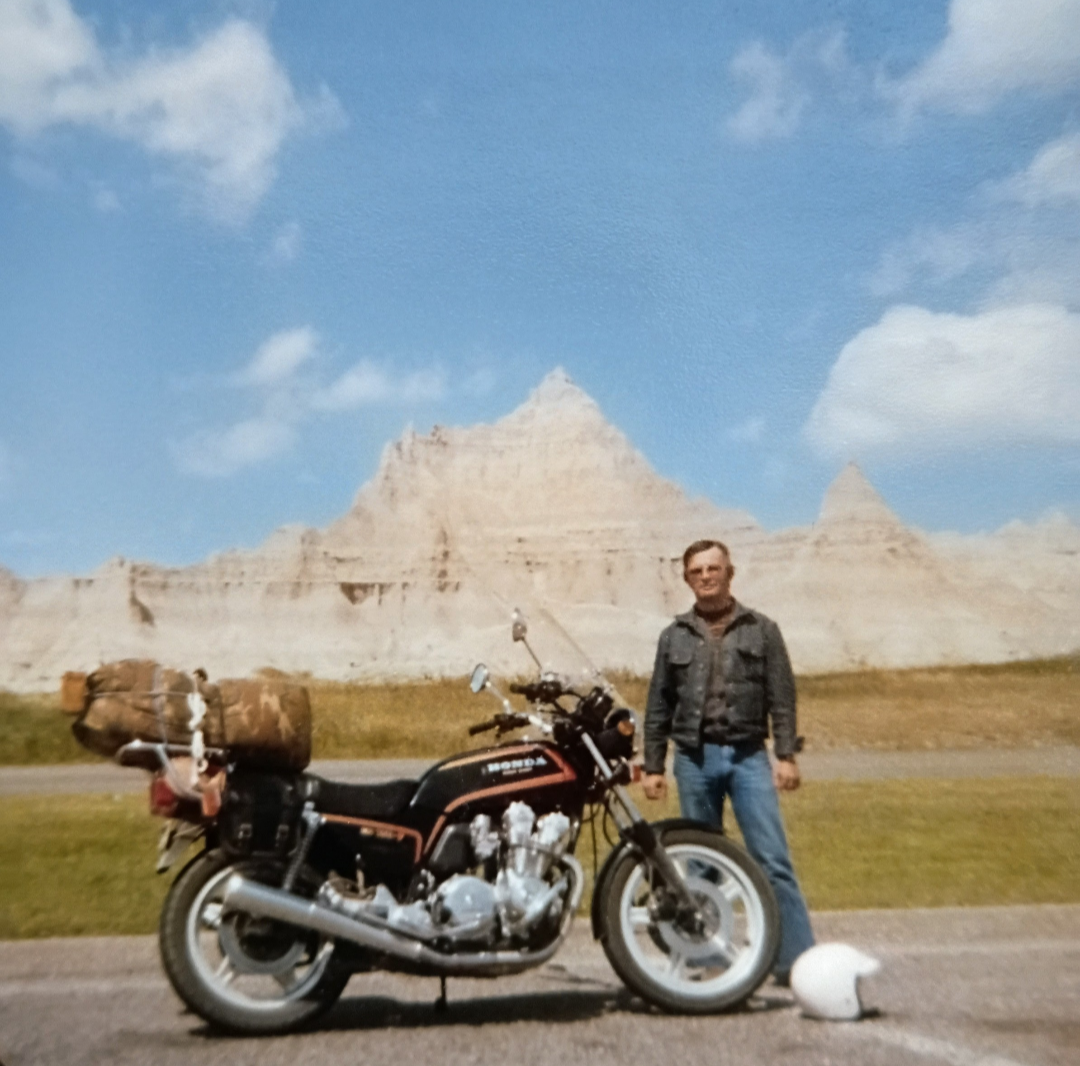 A man standing next to a motorcycle at Mount Rushmore with a white helmet on the ground and blue sky with clouds above.