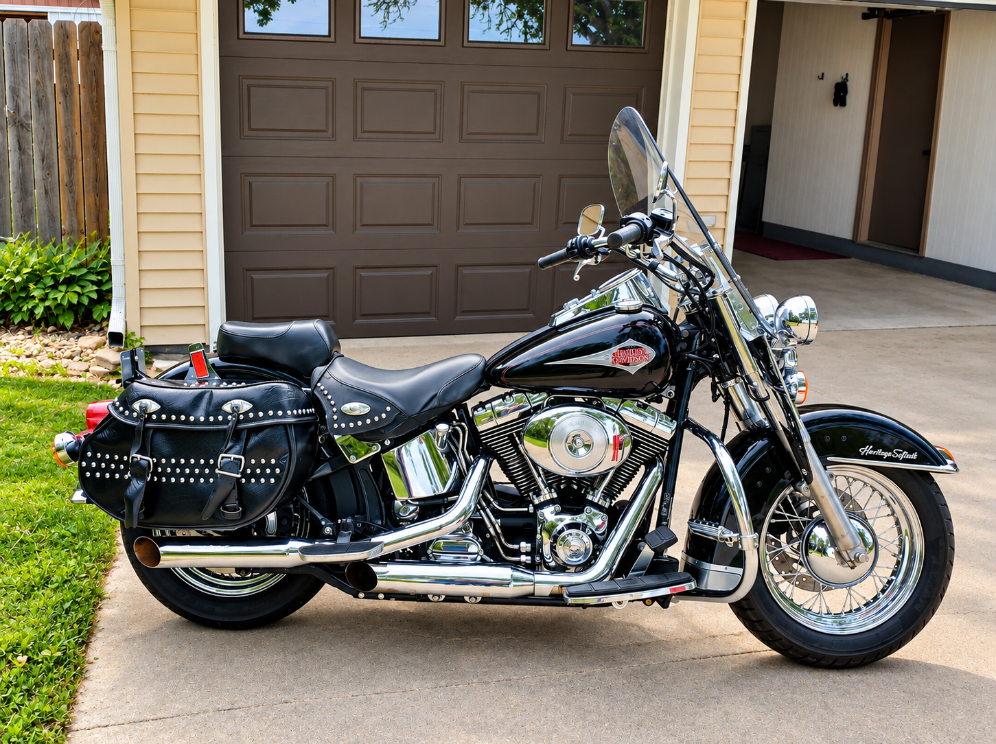 Black Harley-Davidson touring motorcycle with leather saddlebags parked on driveway in front of a garage with a brown door, yellow house siding, and a small garden on the side.