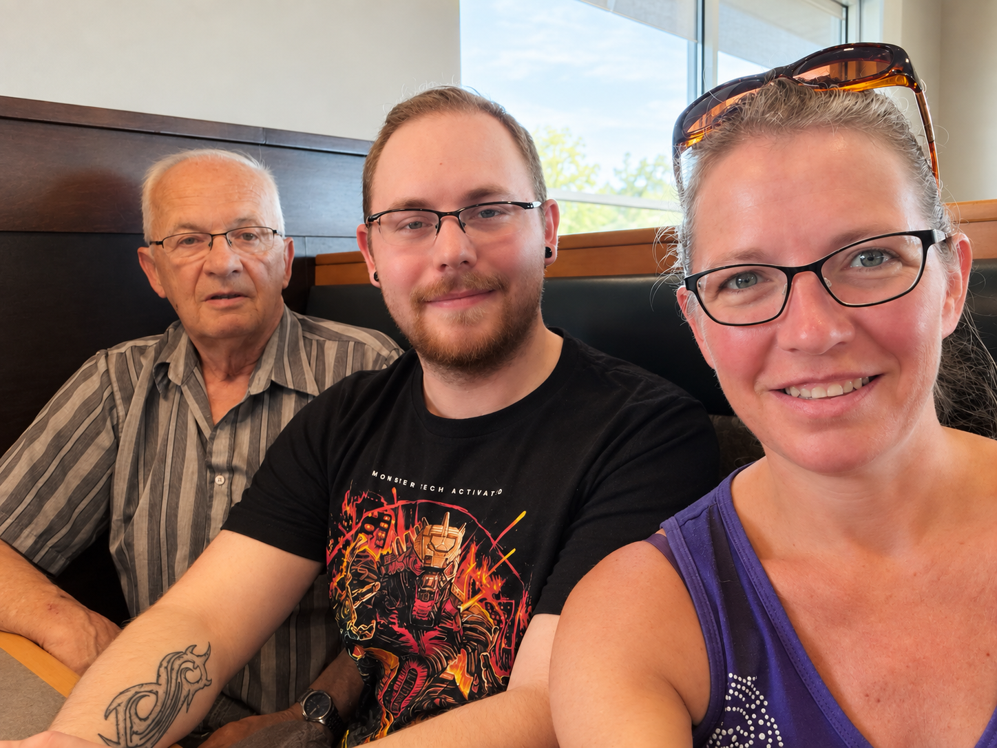 Three people sitting in a restaurant booth, smiling and taking a selfie, with a window showing trees in the background.