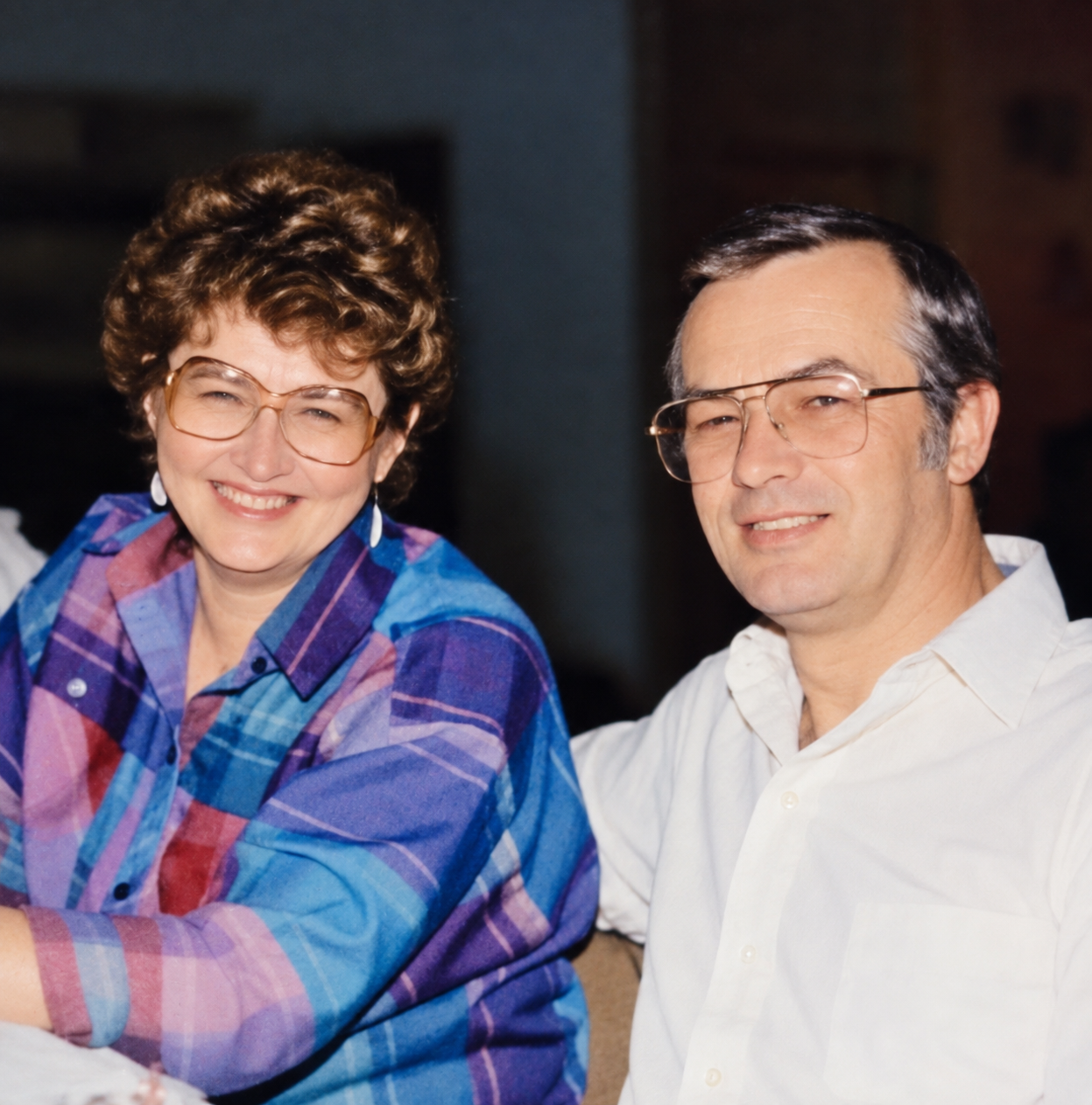 A smiling woman with short curly brown hair wearing large glasses and a colorful plaid shirt, sitting next to a smiling man with dark hair, glasses, and a white collared shirt.