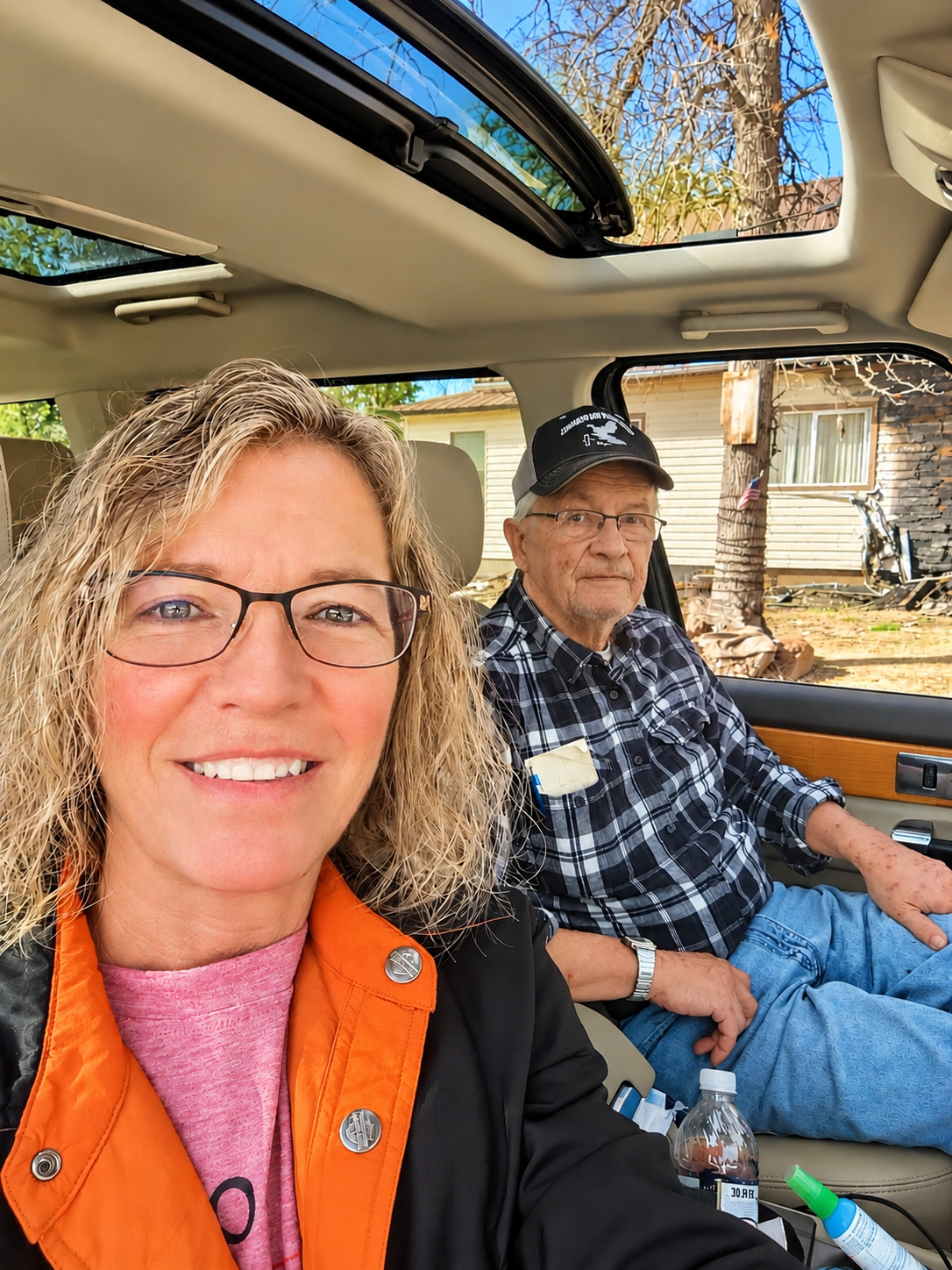 A woman with blonde curly hair and glasses smiling for a selfie inside a vehicle, with an elderly man with a cap and glasses sitting in the background beside a window showing a damaged house and fallen tree outside.