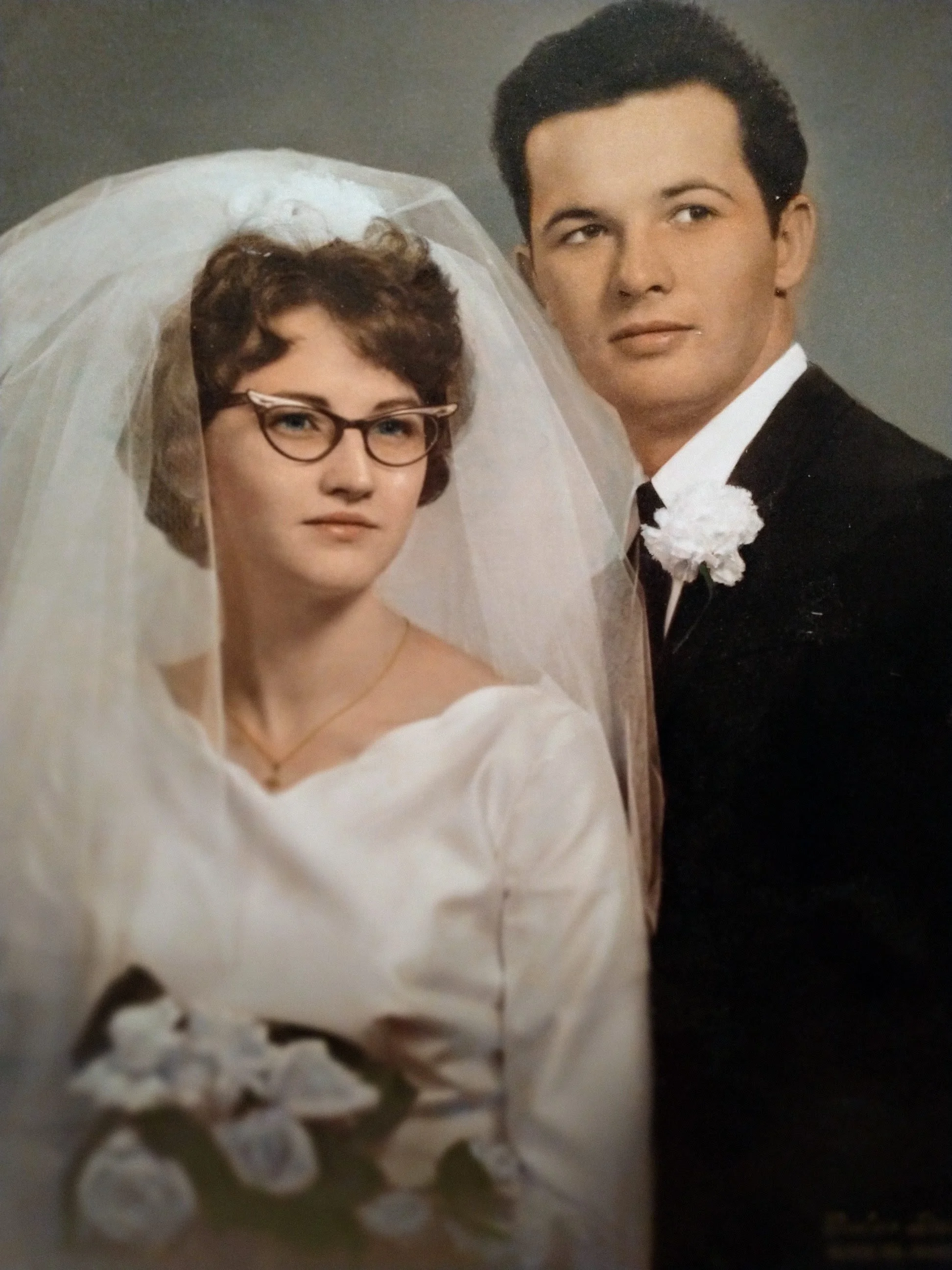 A man and woman dressed in wedding attire, with the woman wearing glasses and a veil, appears in a portrait against a plain background.