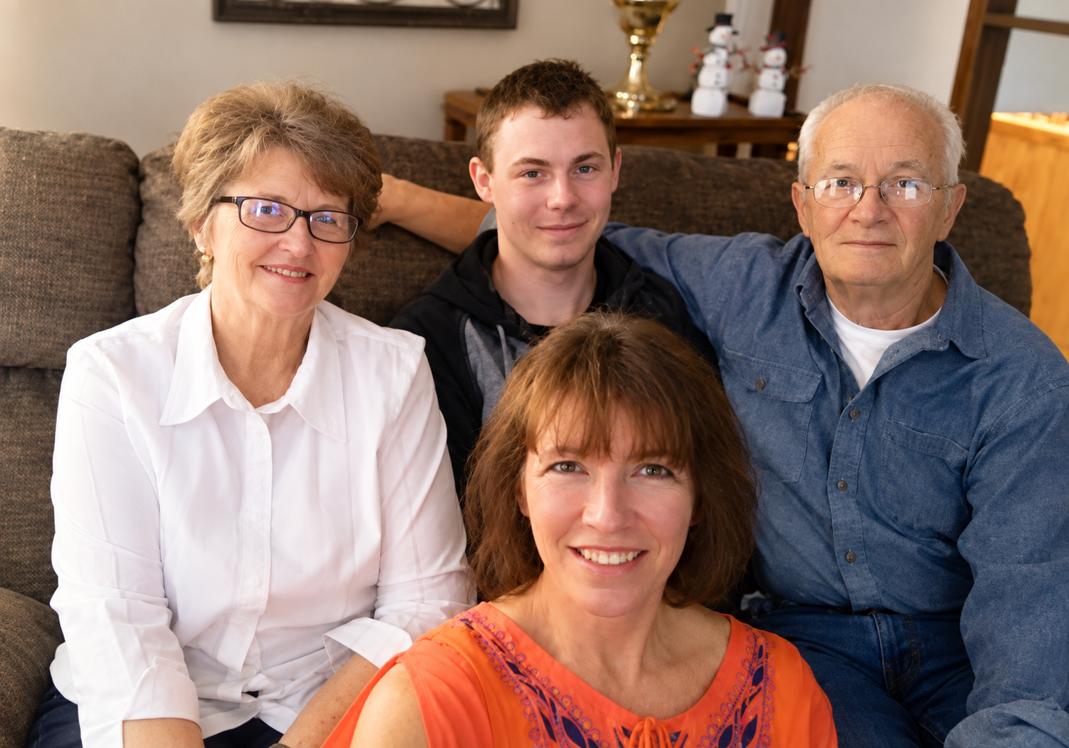 A multi-generational family gathering sitting on a brown couch with holiday decorations in the background, including a small snowman and a golden trophy.