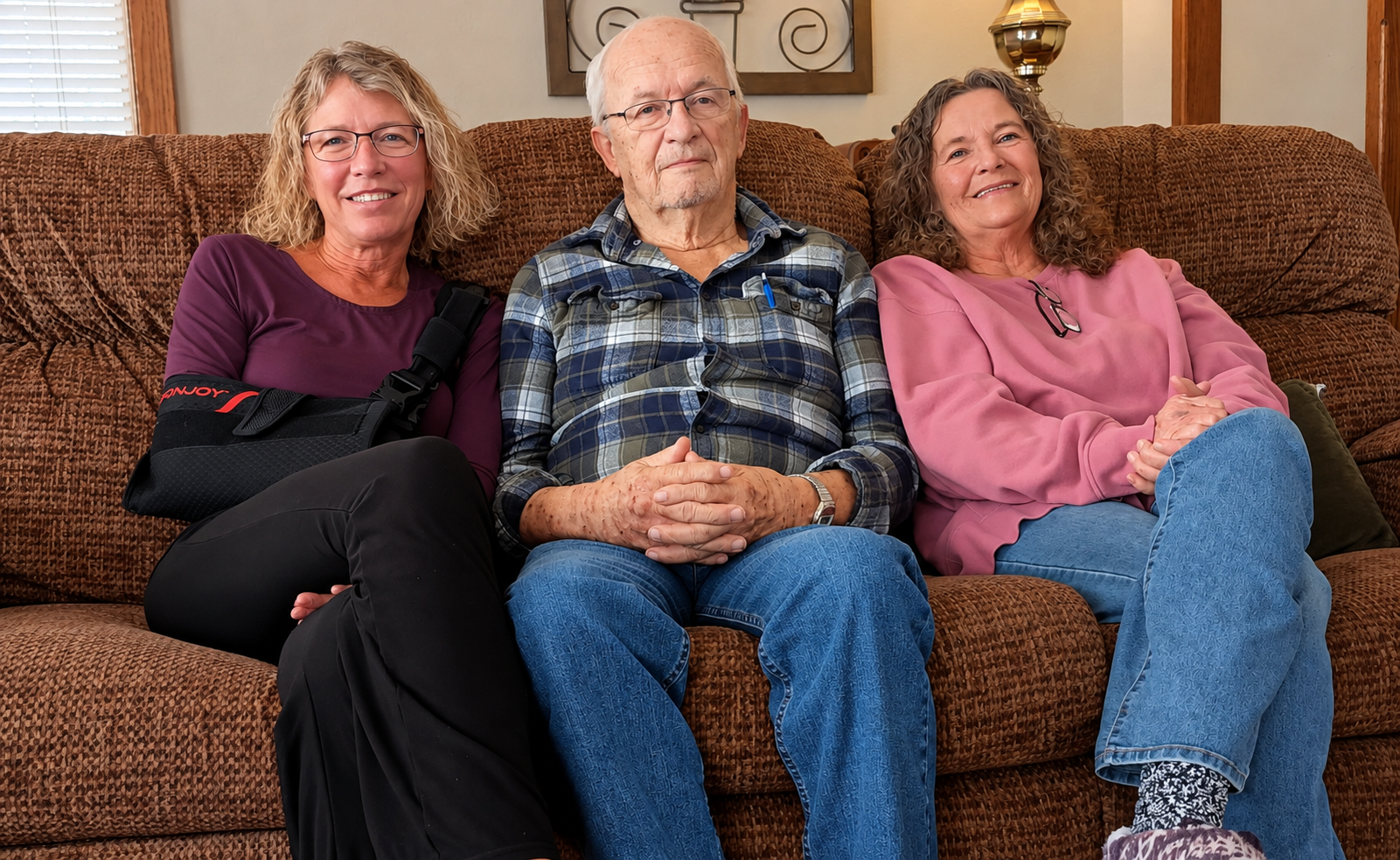 Three elderly people sitting on a brown couch in a living room. The man in the middle wears glasses and a blue plaid shirt, with hands clasped on his lap. The woman on the left has glasses, curly blonde hair, and is wearing a purple top and black pants, with a black arm sling. The woman on the right has curly hair, wears a pink sweatshirt and blue jeans, with glasses hanging around her neck. All are smiling or looking content.