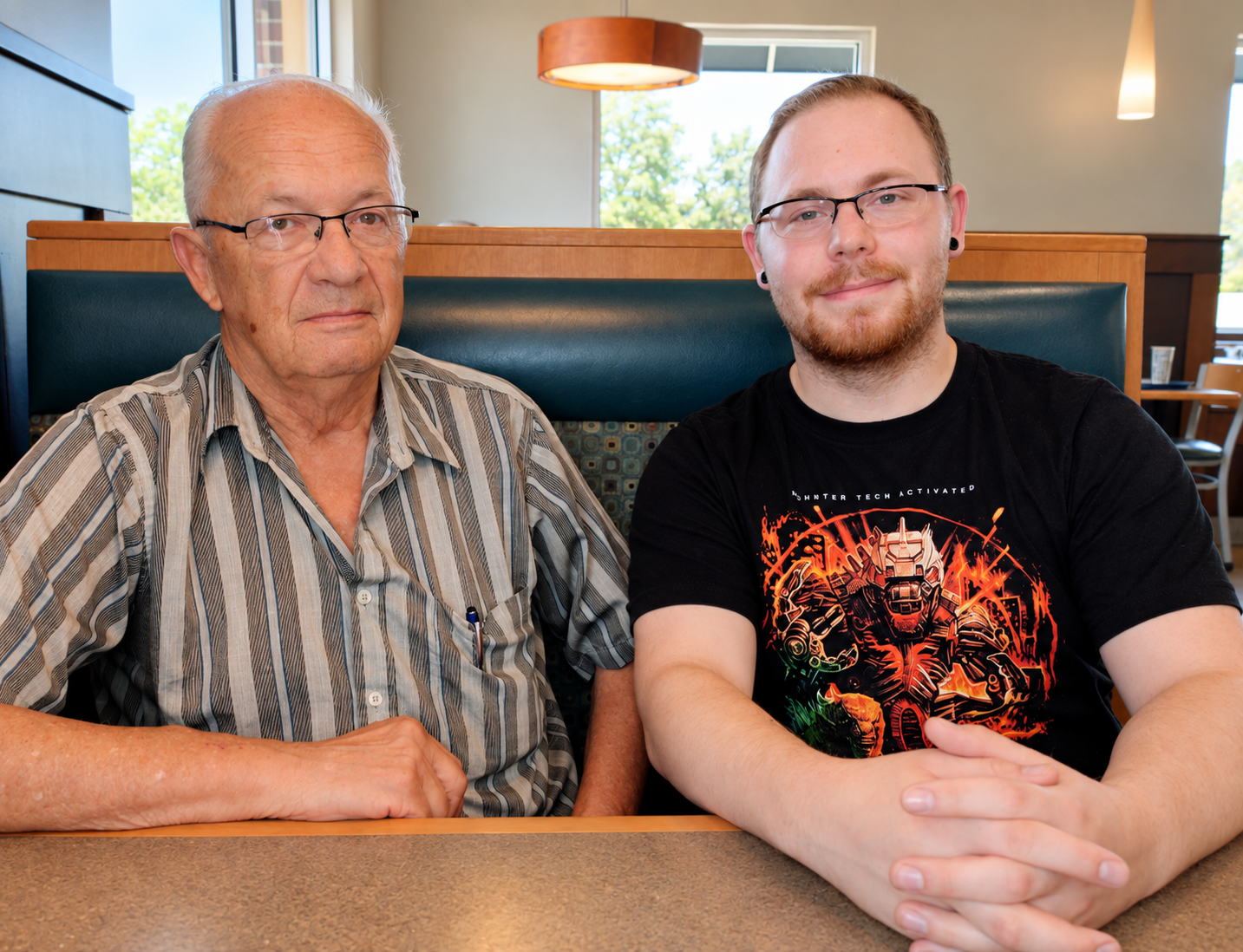 An elderly man and a young man sit side by side at a restaurant booth, both smiling slightly and facing the camera.