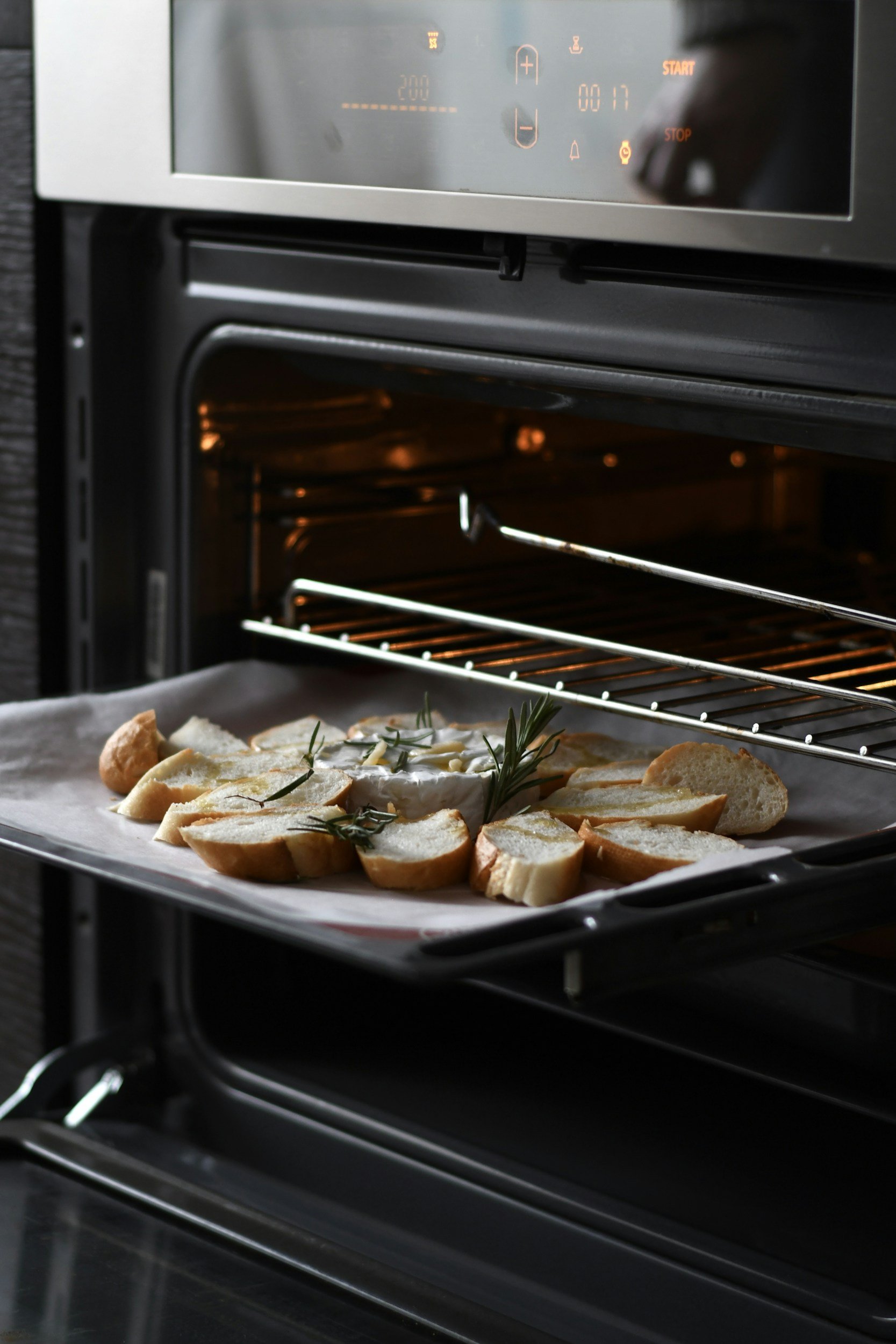 An oven with the door open, revealing bread slices on a baking tray inside, and the oven's digital display showing temperature and settings.