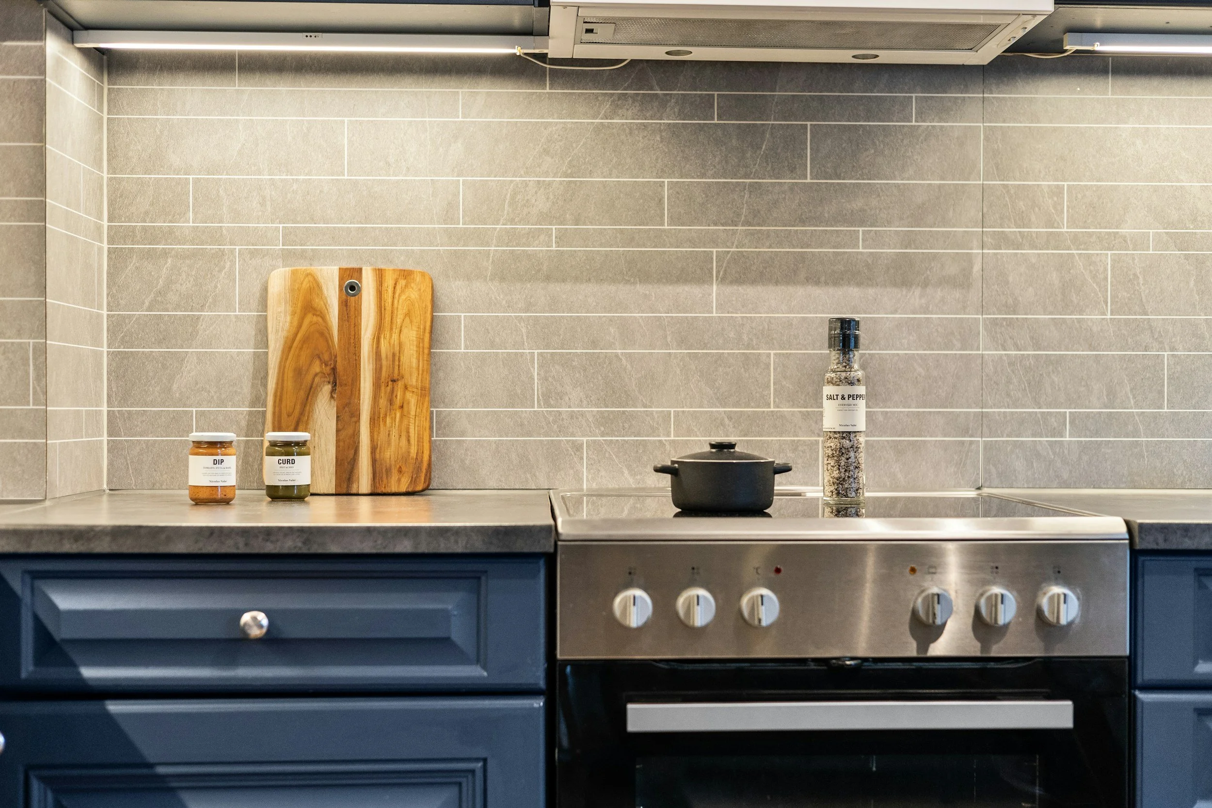 Kitchen with gray tile backsplash, blue cabinetry, and a stainless steel stove. On the countertop are jars labeled 'DIP', 'CURD', and 'SALT & PEPPER', a wooden cutting board, and a black pot on the stove.