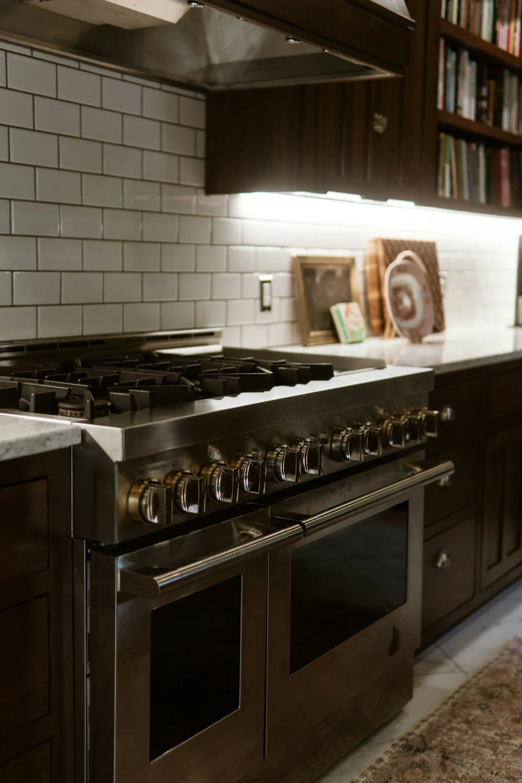Stainless steel kitchen stove with oven and black knobs, white tile backsplash, wooden upper cabinets, books and framed pictures on the counter, and a patterned rug on the floor.