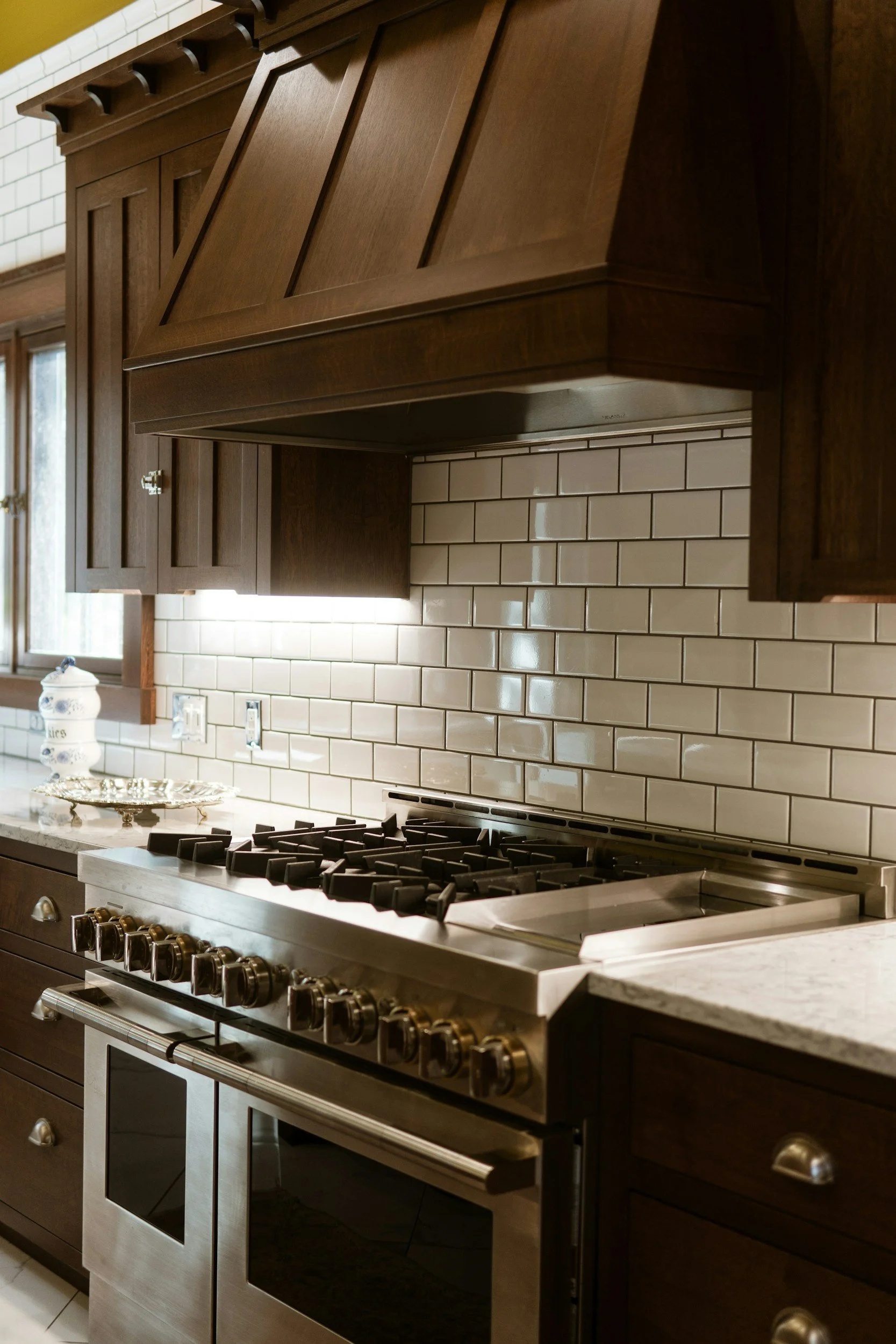 A modern kitchen featuring white subway tile backsplash, a stainless steel gas range oven, and wooden cabinets with a dark brown finish.