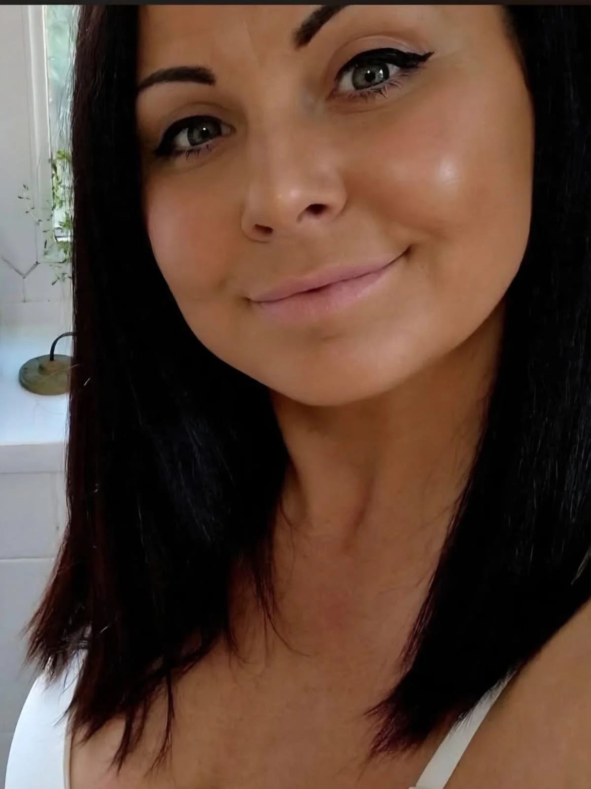 A close-up of a woman with black hair and gray eyes, smiling softly, with winged eyeliner and light lipstick, in a well-lit room.