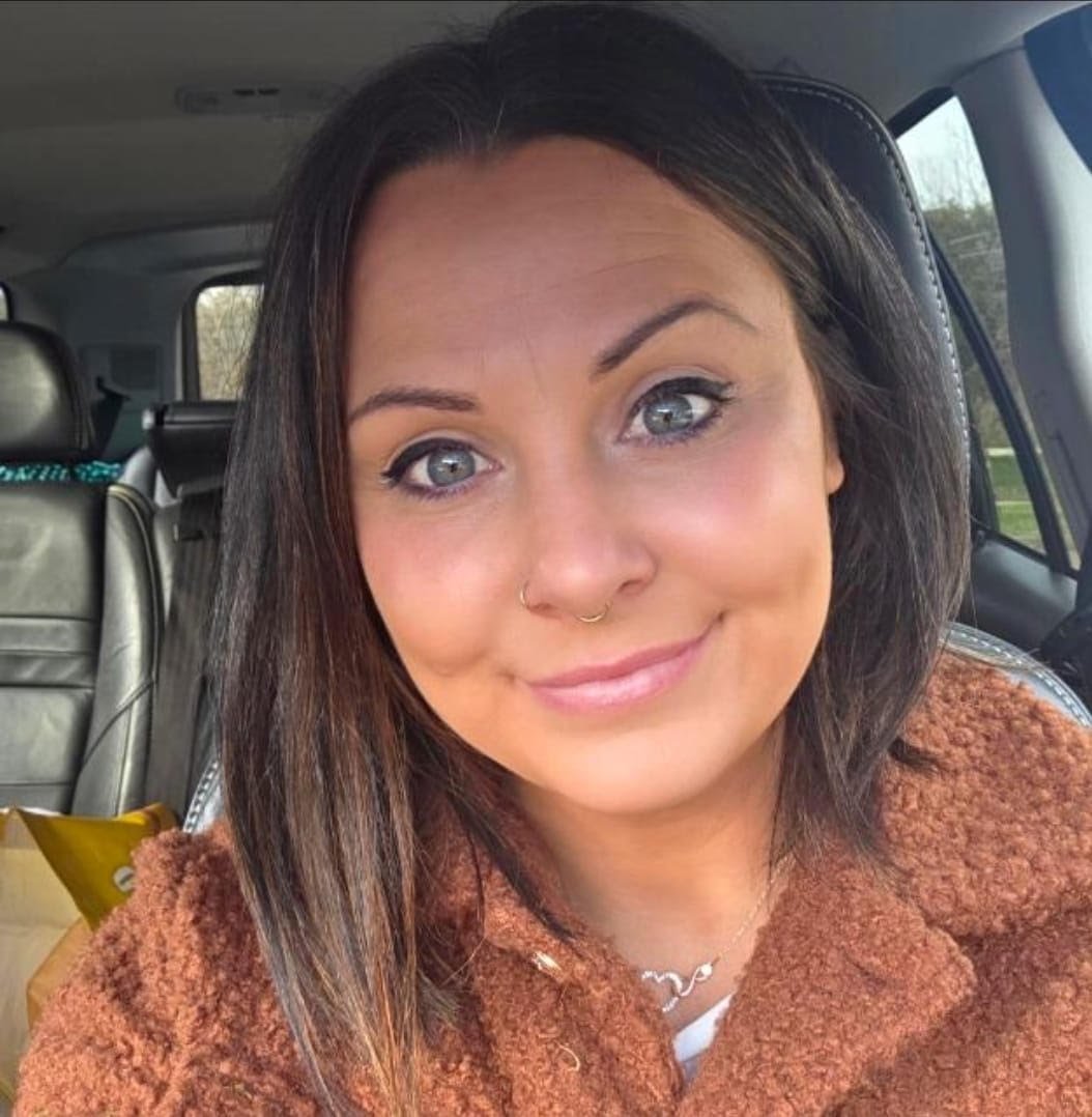 A woman with dark brown hair and blue eyes smiling inside a car, wearing a brown jacket and a silver necklace.