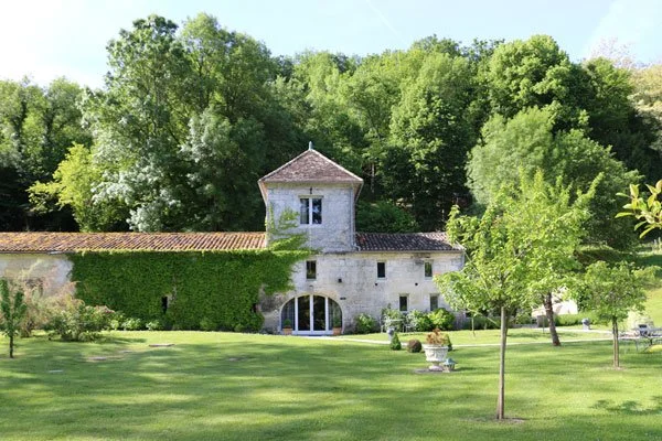 A stone house with a small tower surrounded by a well-maintained lawn and several trees, with a forested hillside in the background.