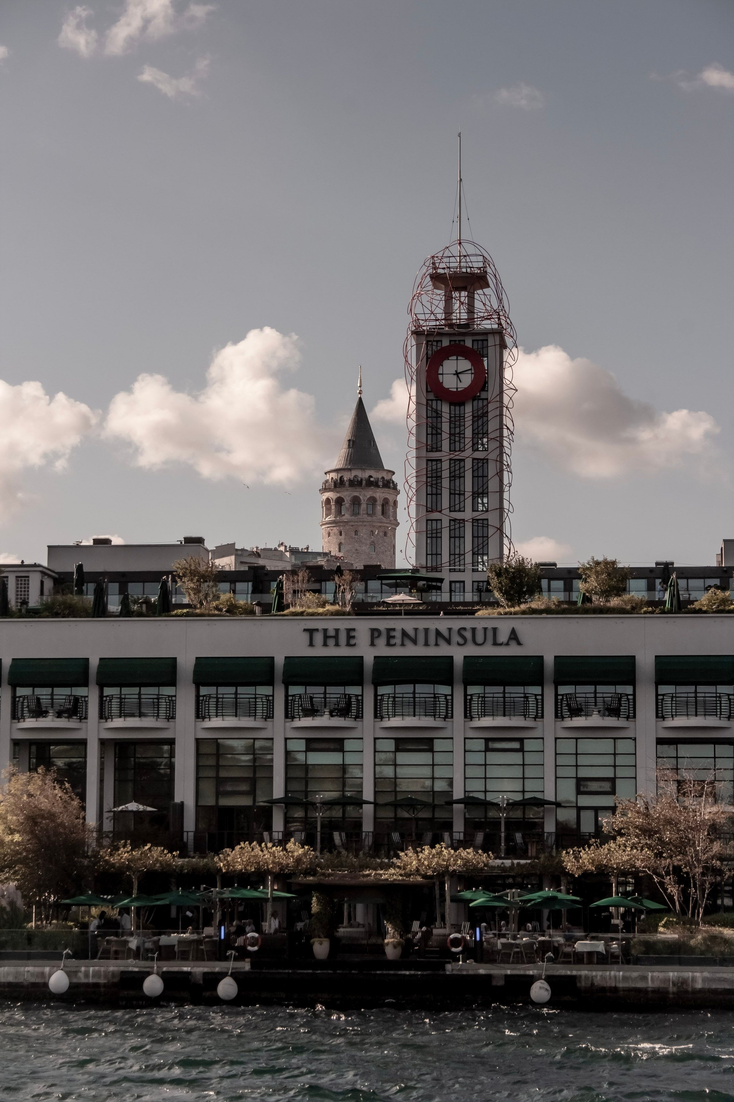 A building labeled 'The Peninsula' with a skyline featuring the Galata Tower in Istanbul, Turkey, visible in the background. The scene is taken from across a body of water with structures along the water's edge.
