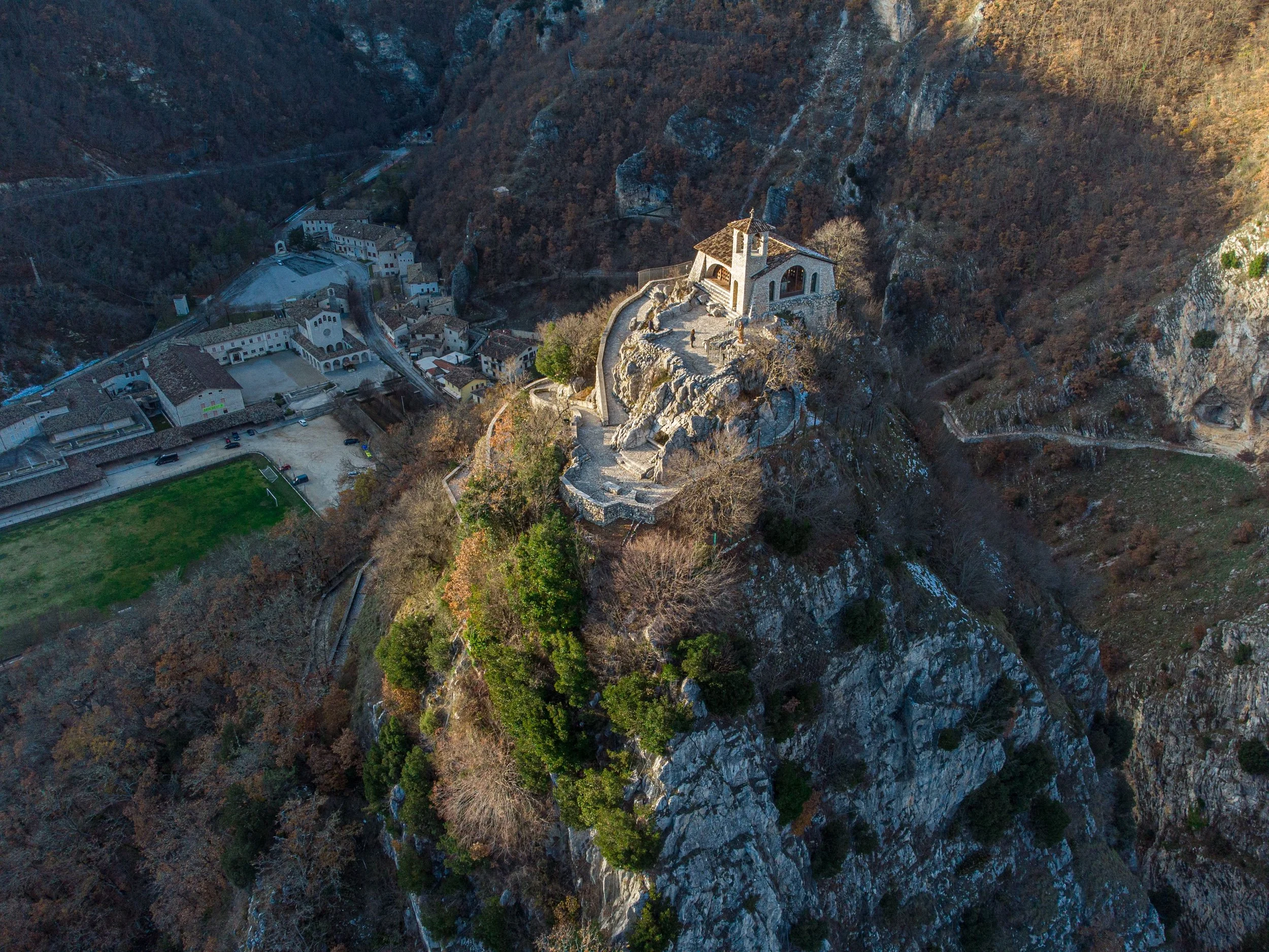 An aerial view of a mountainous area with a small village at the base and a church-like building perched on a rocky hilltop, connected by a winding pathway.