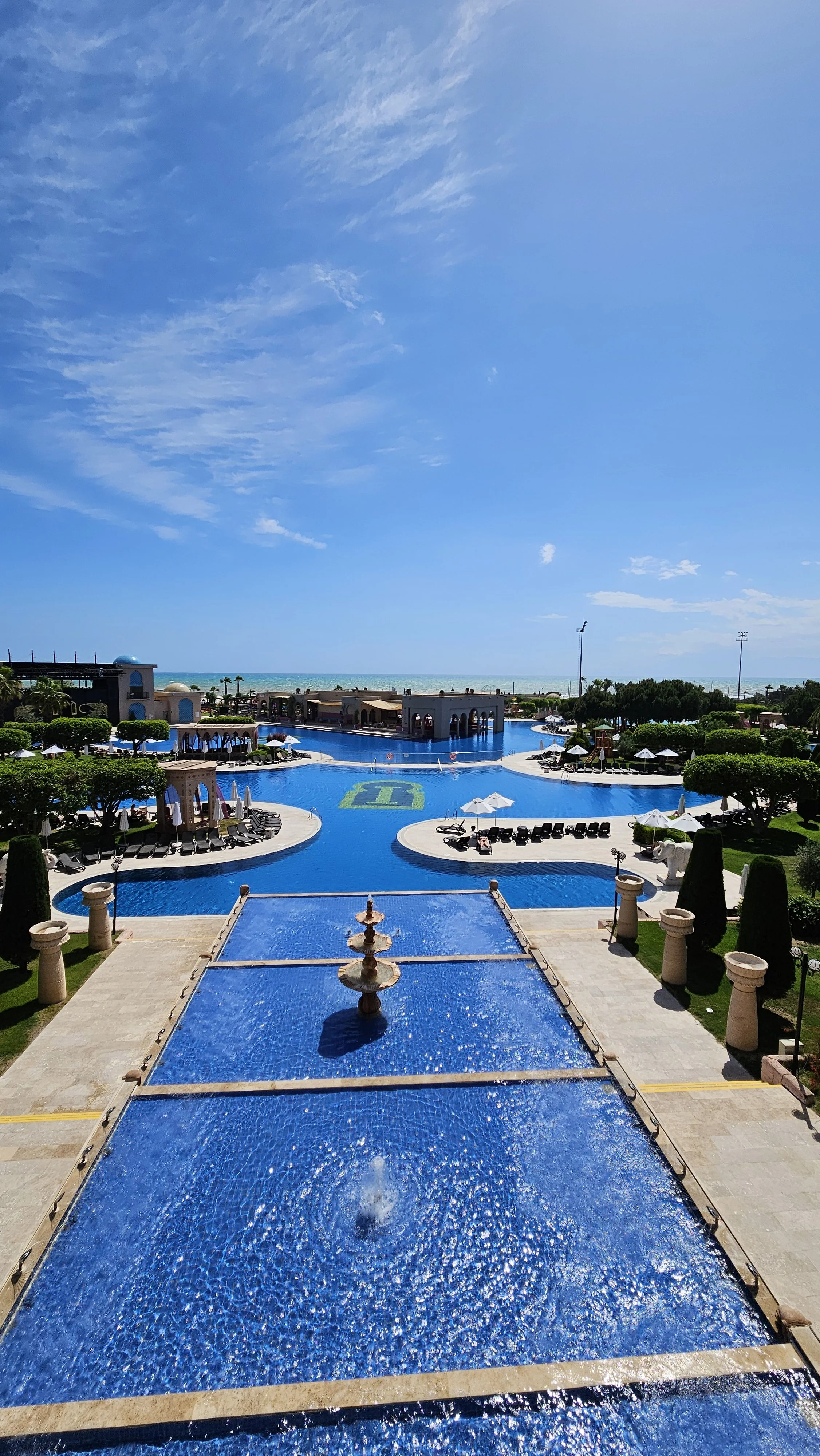 Resort pool area with a large blue swimming pool, lounge chairs with umbrellas, manicured trees, and a view of the ocean in the background.