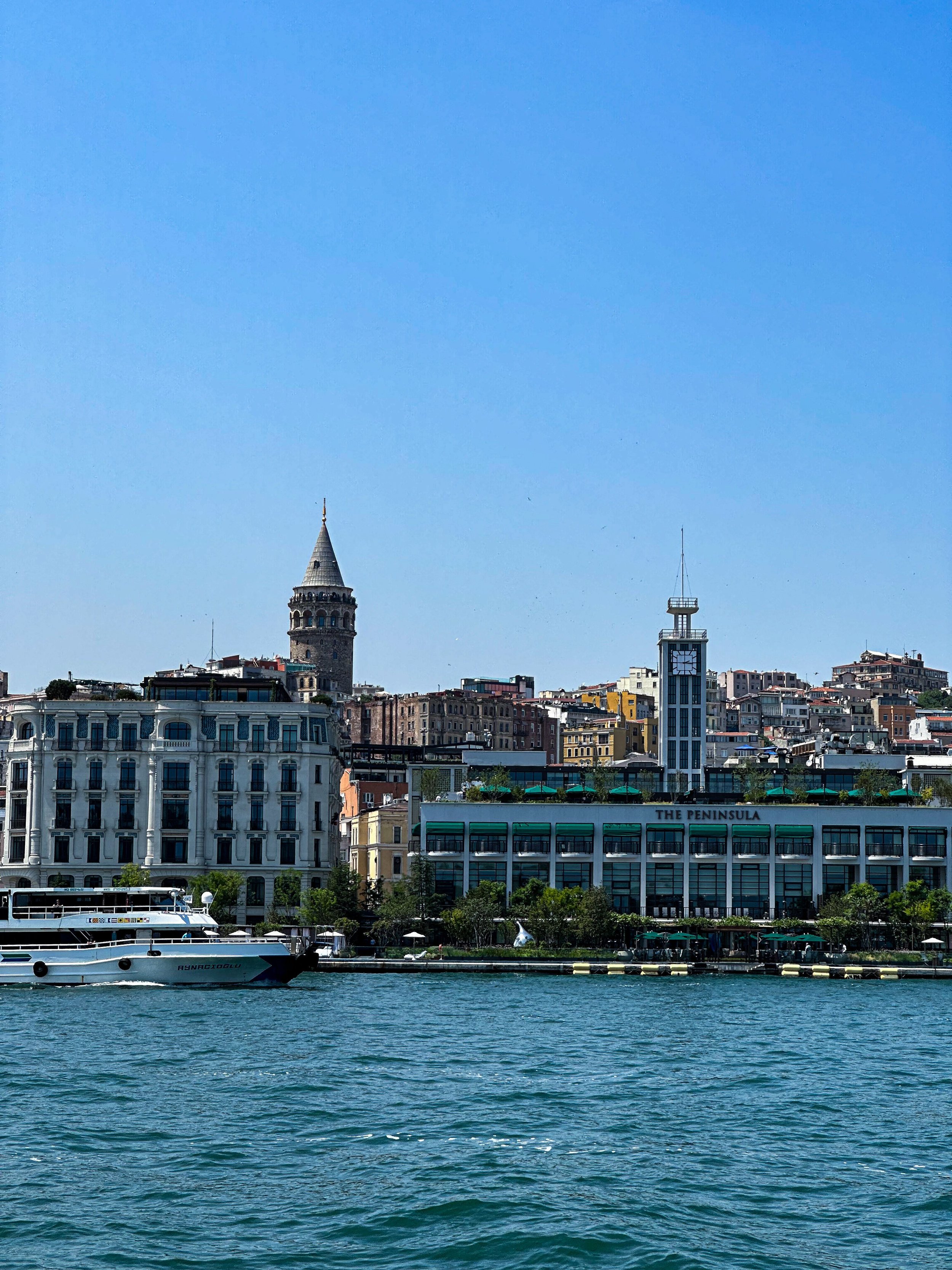 Cityscape with historic tower and modern buildings along waterfront, boat in foreground, clear blue sky