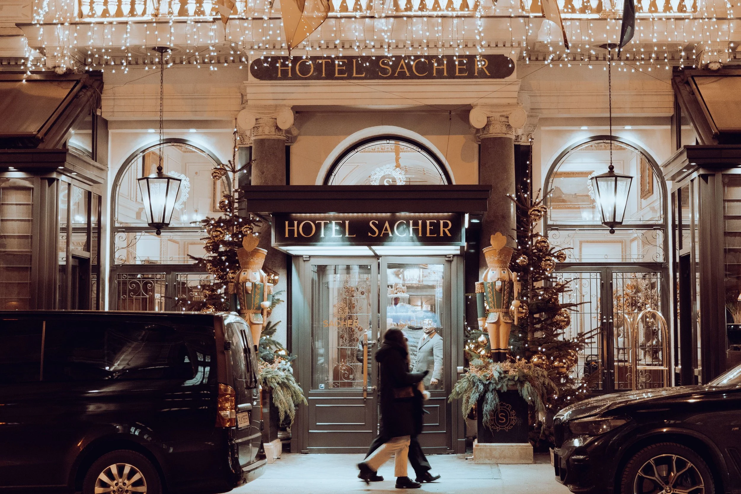 Nighttime scene of the entrance to Hotel Sacher decorated with Christmas lights, wreaths, and ornaments, with people walking past and cars parked outside.