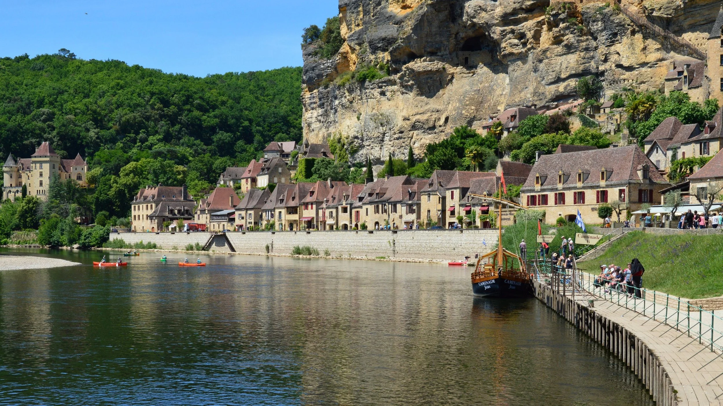 A riverside village with historic stone houses and a large cliff behind, people are leisurely walking and kayaking on the river, with a decorative boat docked at a pier.