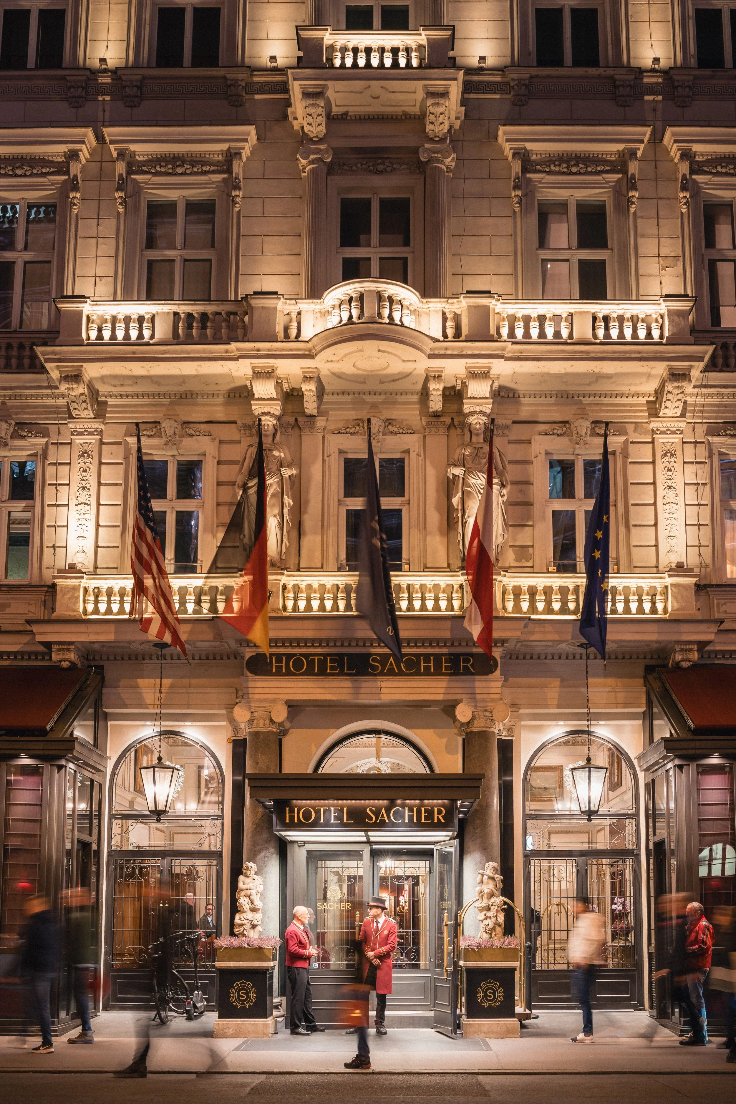 Exterior of the Hotel Sacher, a grand historic building illuminated at night with decorative statues and multiple flags, including the American and European Union flags, at the entrance and people walking by.