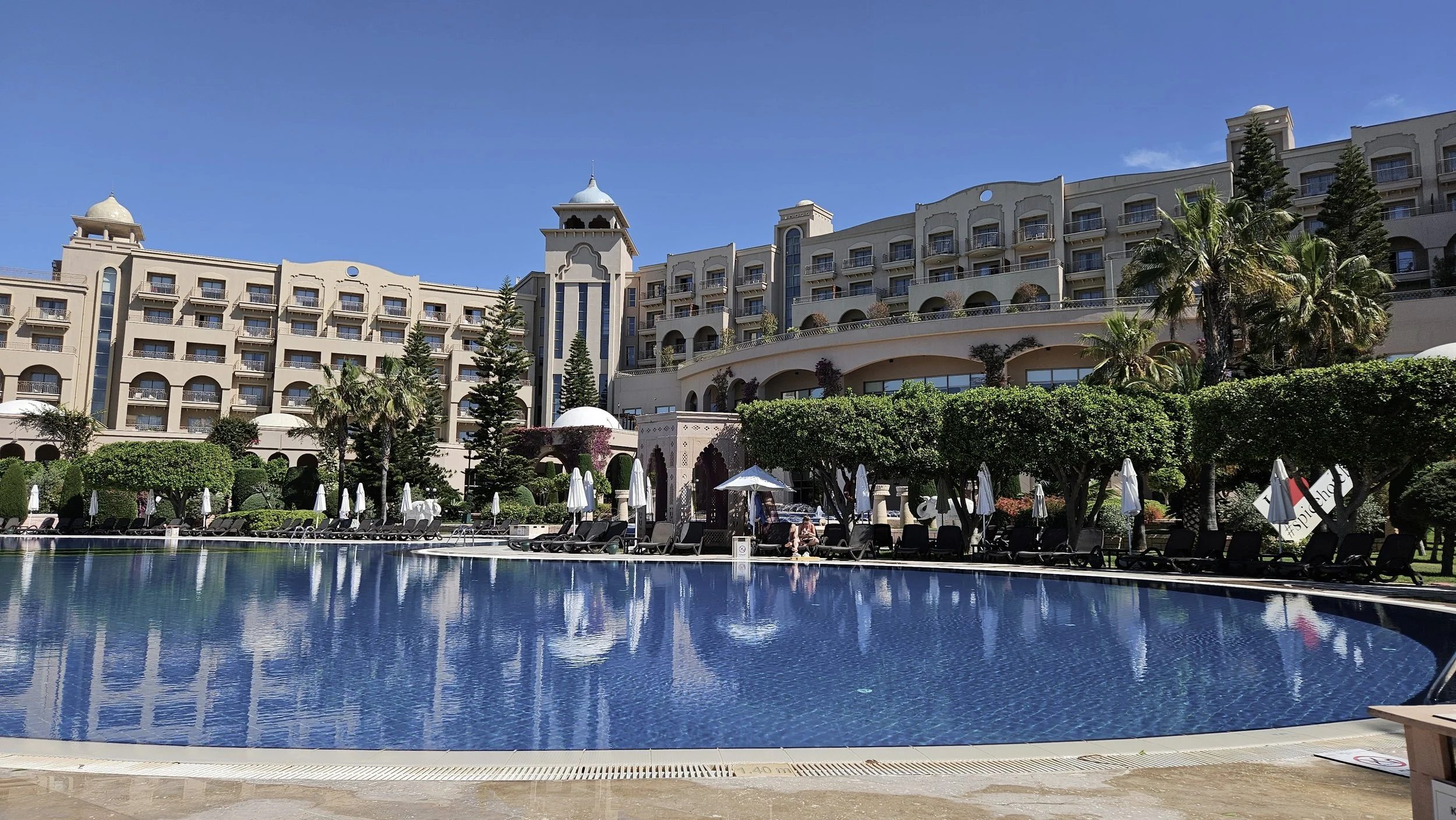 A large hotel or resort building with multiple floors, balconies, and towers, surrounded by greenery, and a swimming pool in the foreground with lounge chairs and umbrellas.