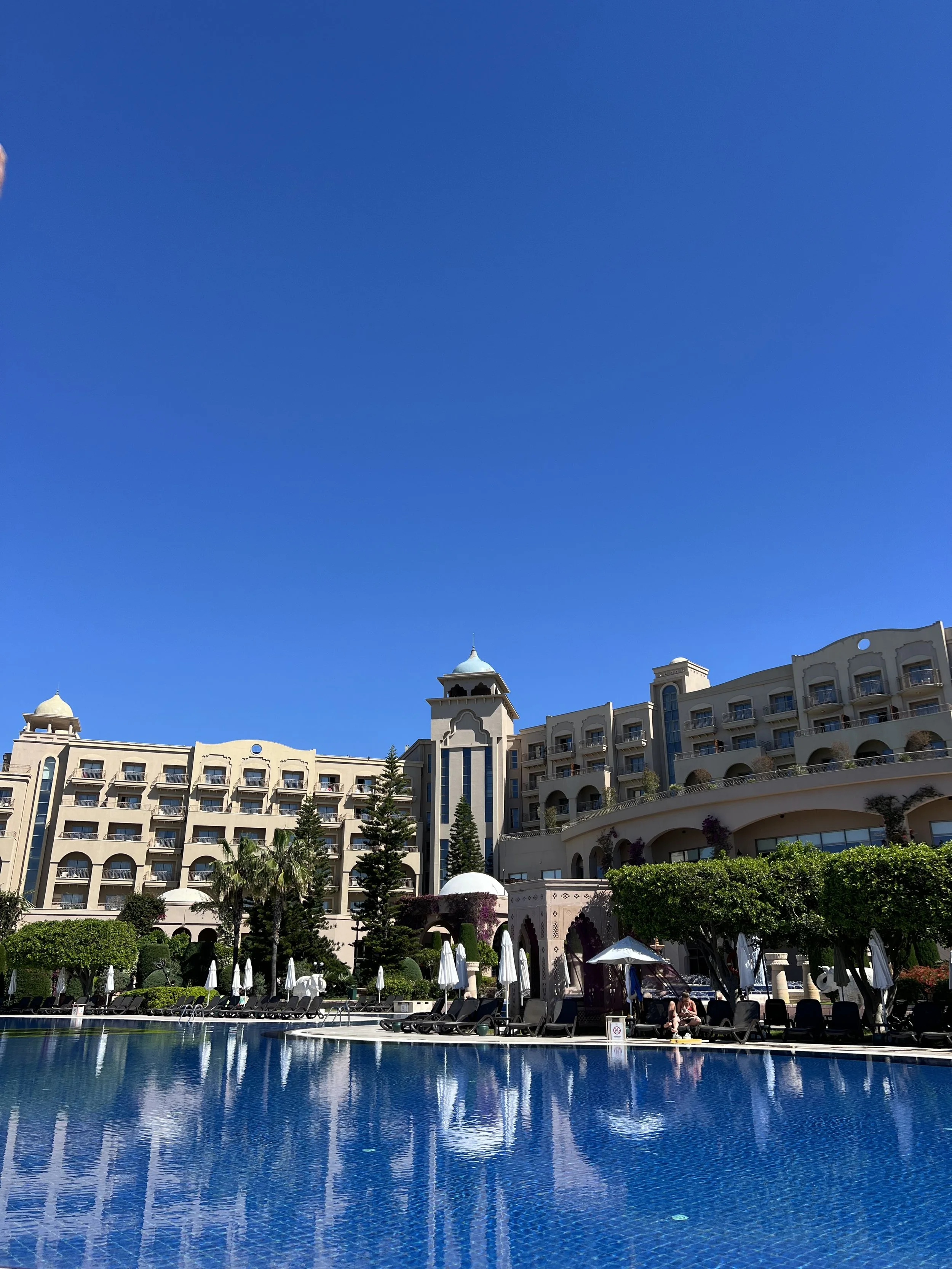 Resort hotel with a large swimming pool, surrounded by lounge chairs and umbrellas, with a multi-story building featuring towers and balconies, and lush greenery against a bright blue sky.