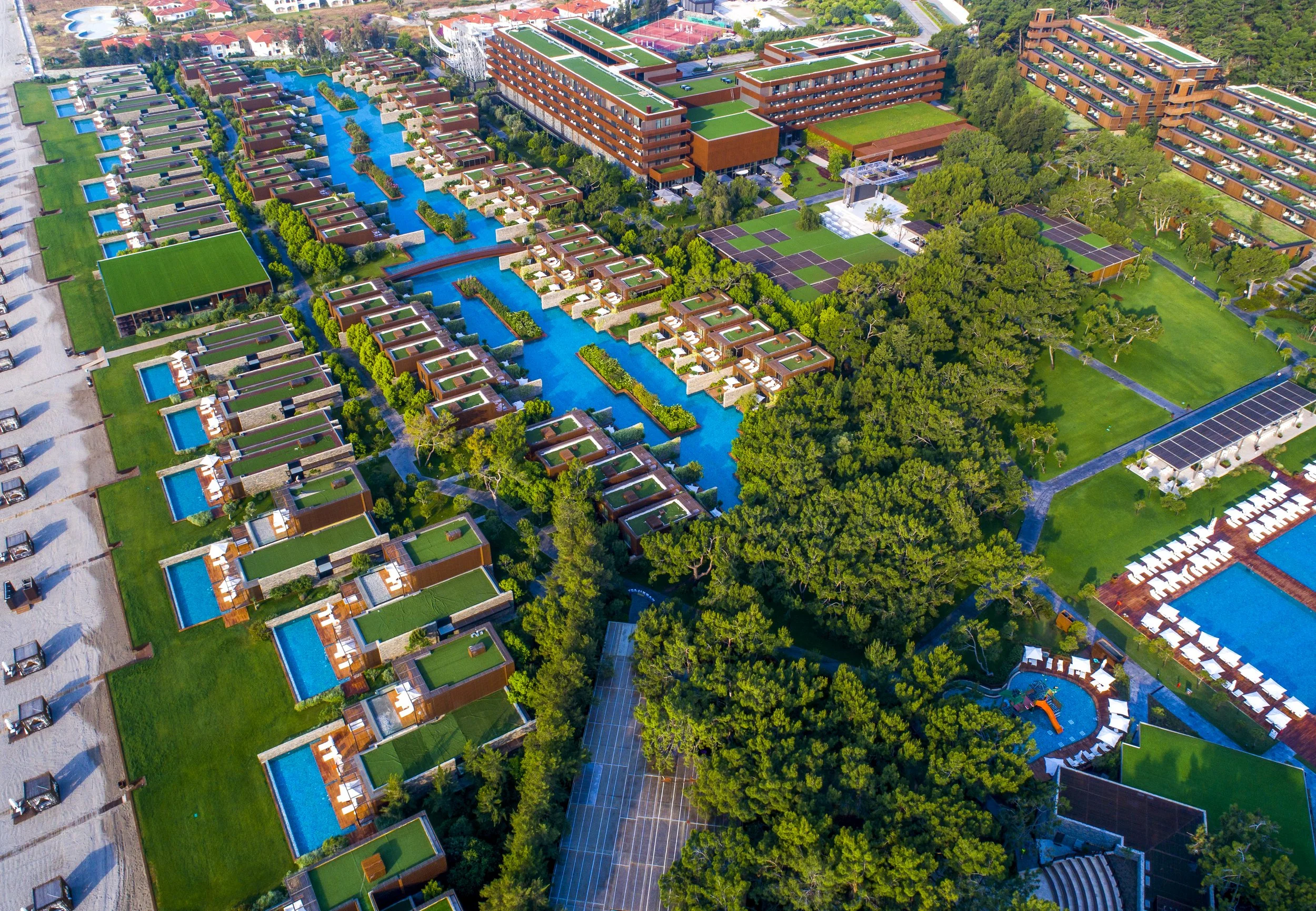 Aerial view of a large resort complex with multiple buildings, swimming pools, water channels, green lawns, tennis courts, and surrounding trees.
