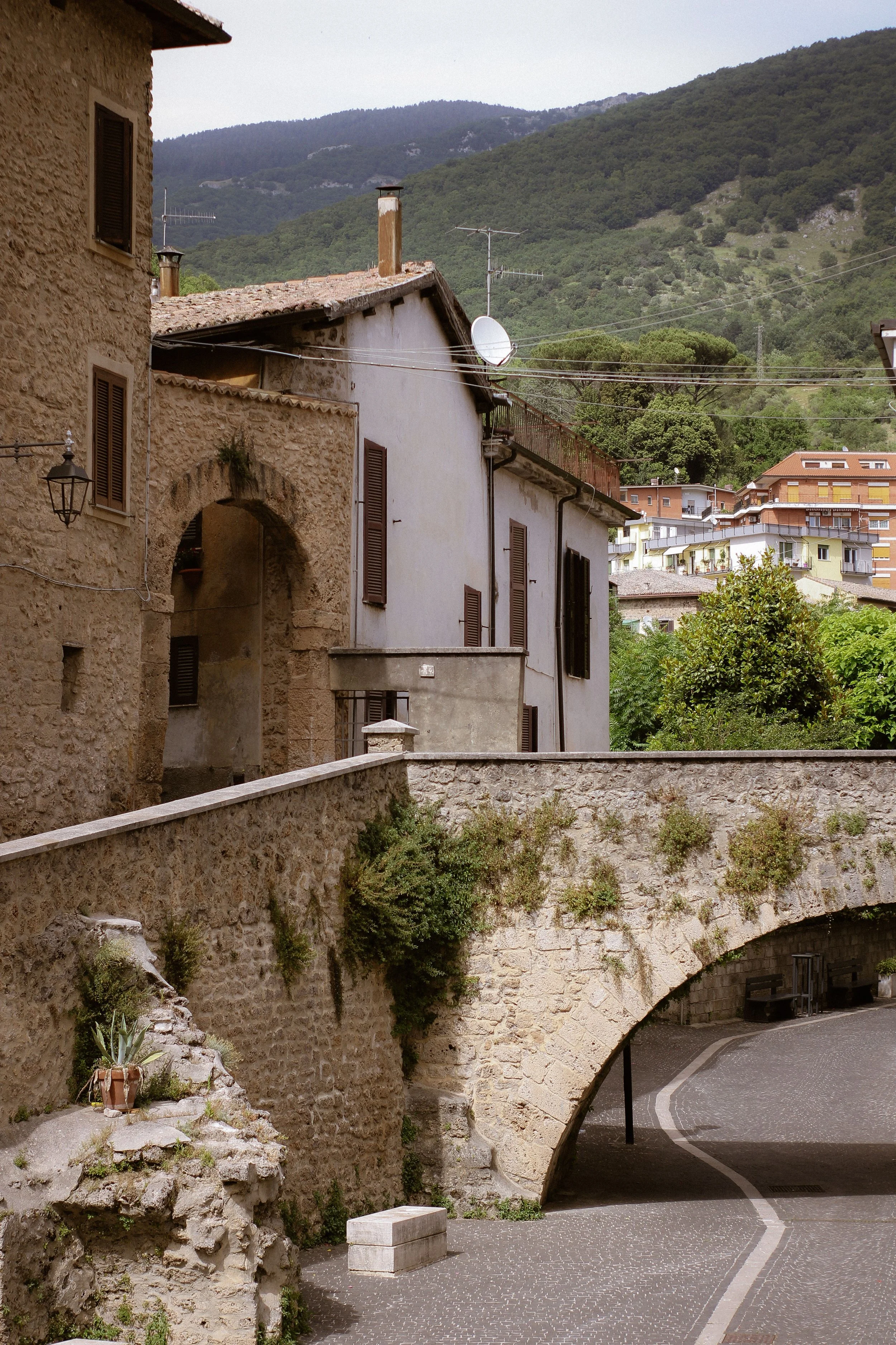 View of a stone bridge with an arch over a curved cobblestone street in a hilly town. The bridge has greenery growing on its side, and there are old stone and stucco buildings with shutters on either side of the street. In the background, lush green mountains and modern apartment buildings are visible.