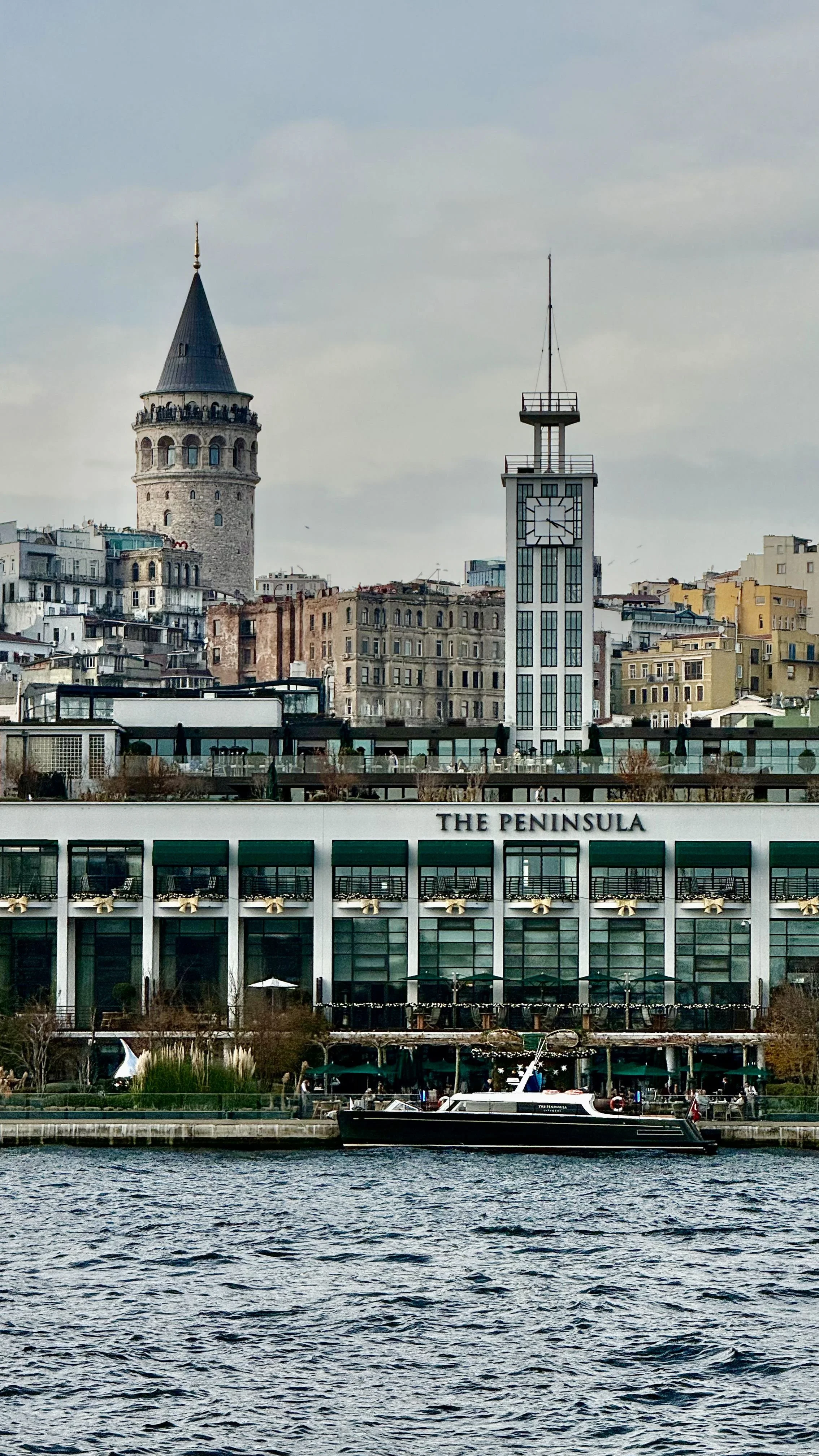 View of Istanbul skyline featuring the Galata Tower and a modern clock tower, with a boat on the water in the foreground and a building labeled 'The Peninsula' below.