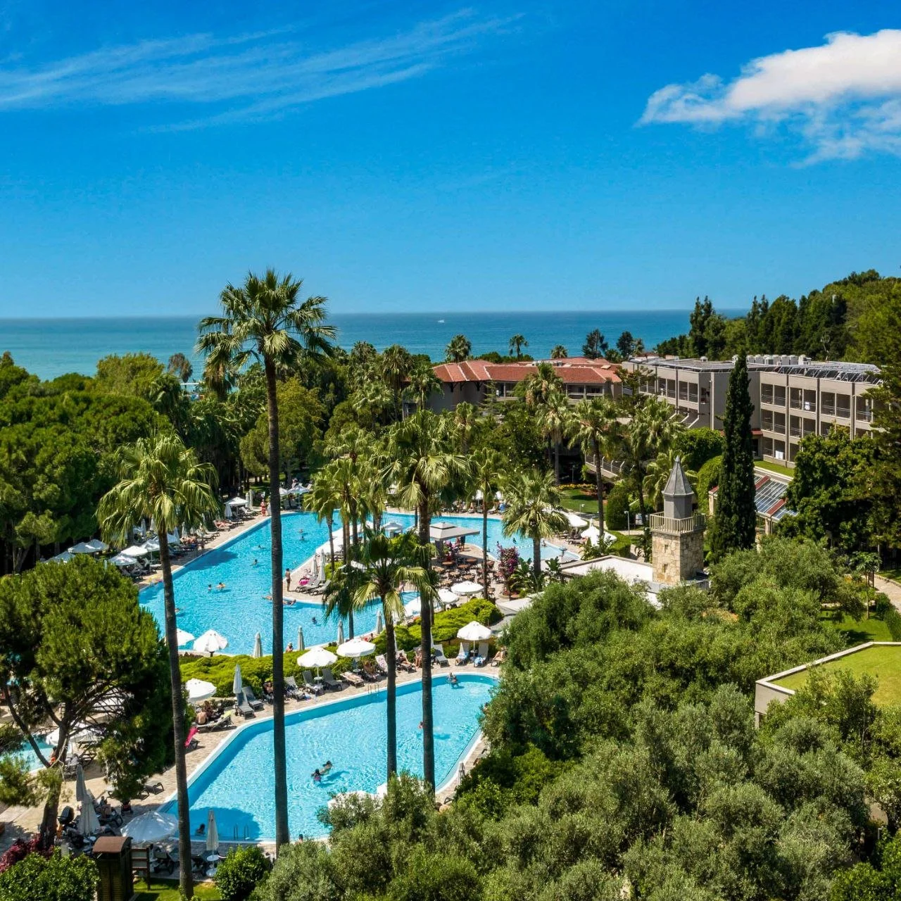 A scenic view of a resort with two swimming pools surrounded by palm trees, umbrellas, and lounge chairs, with a backdrop of the ocean and blue sky with some clouds.