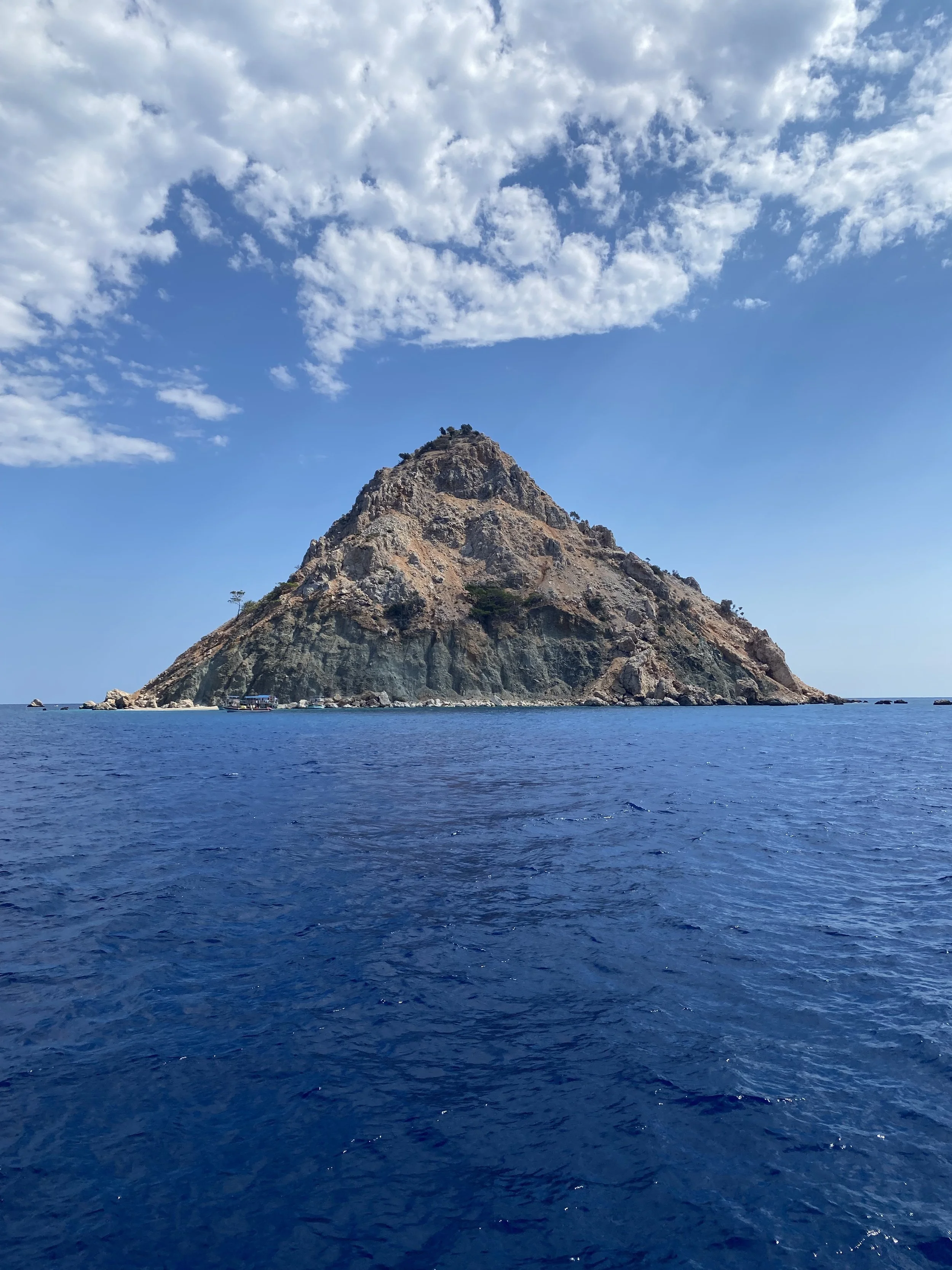 A rocky mountain island surrounded by blue ocean water with small boats near the base, under a partly cloudy sky.