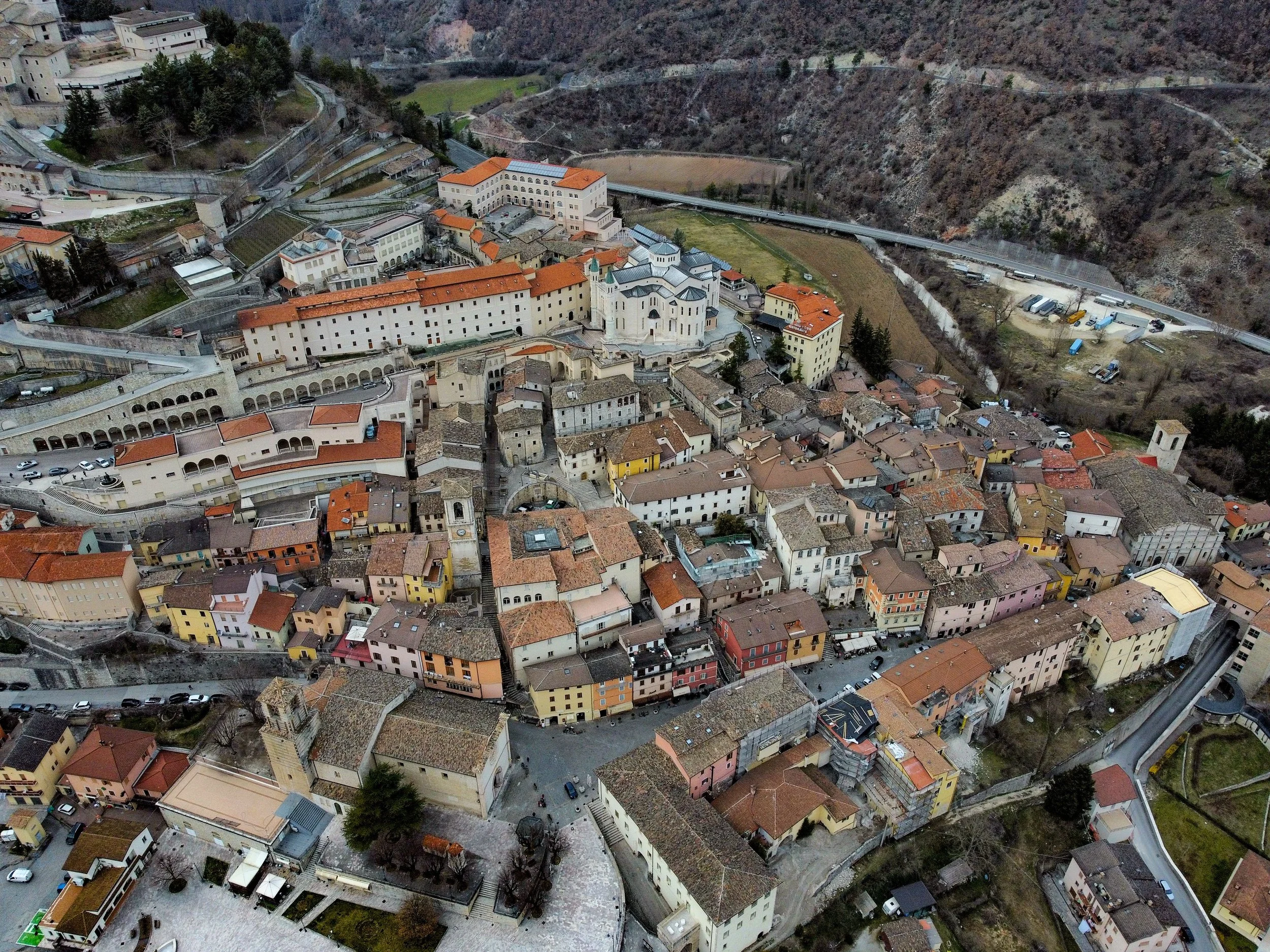 An aerial view of a small European town nestled in a valley surrounded by hills and mountains. The town features a mix of historic buildings with red-tiled roofs, narrow streets, and a prominent church with twin towers. Roads and parking areas are visible along the periphery, and there is a steep hillside with a road winding around it in the background.