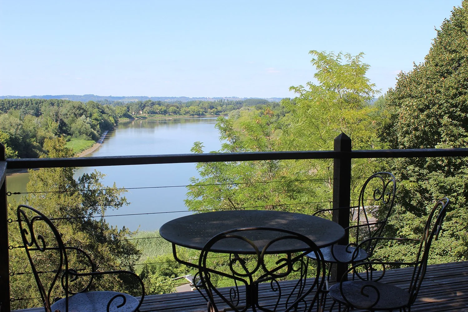 A view of a river with lush green trees on both sides from a balcony with a black metal table and chairs.
