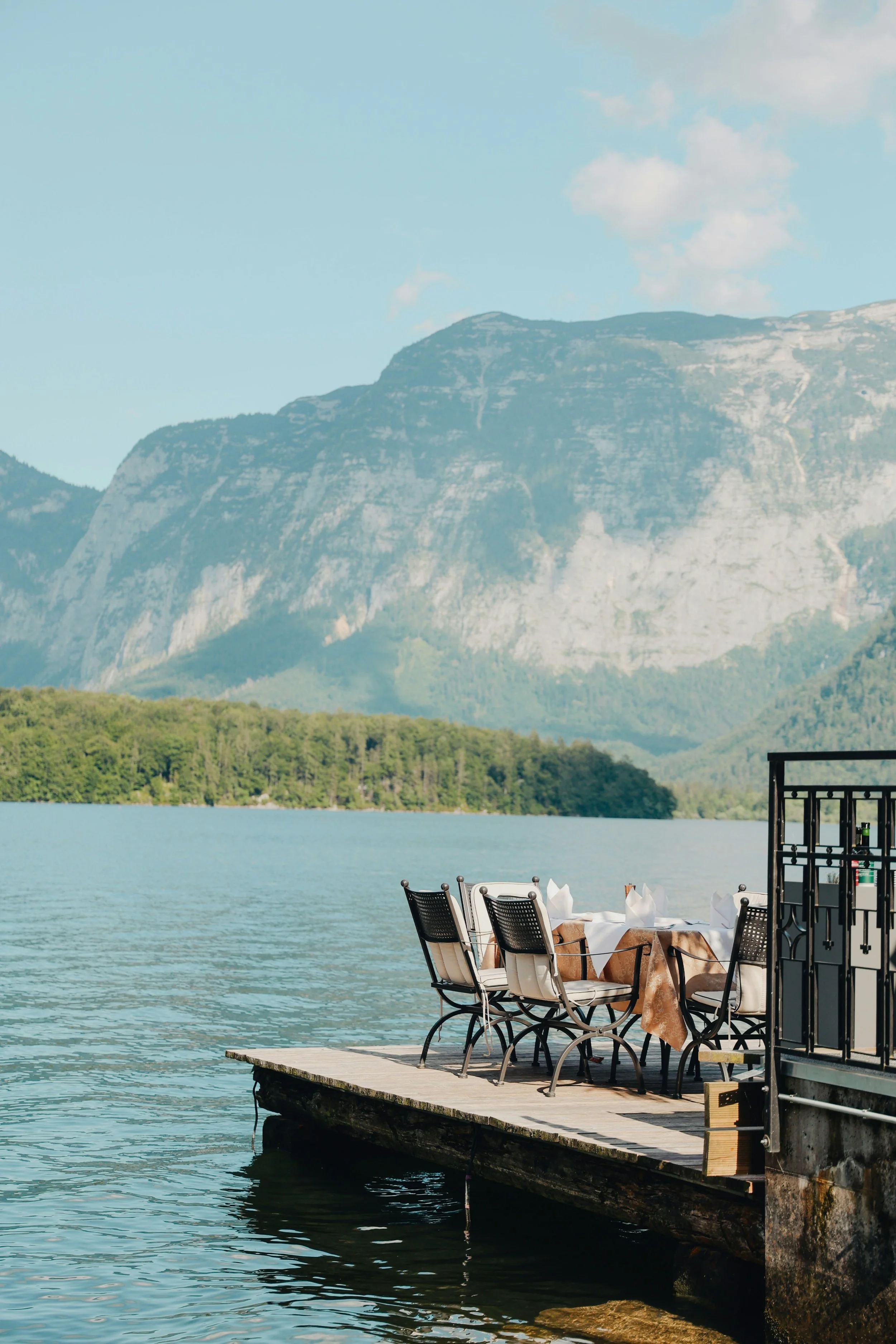 A lakeside outdoor dining area with chairs and a table set with napkins and a tablecloth, situated on a wooden dock next to calm water, with green trees, a mountain range, and a partly cloudy sky in the background.