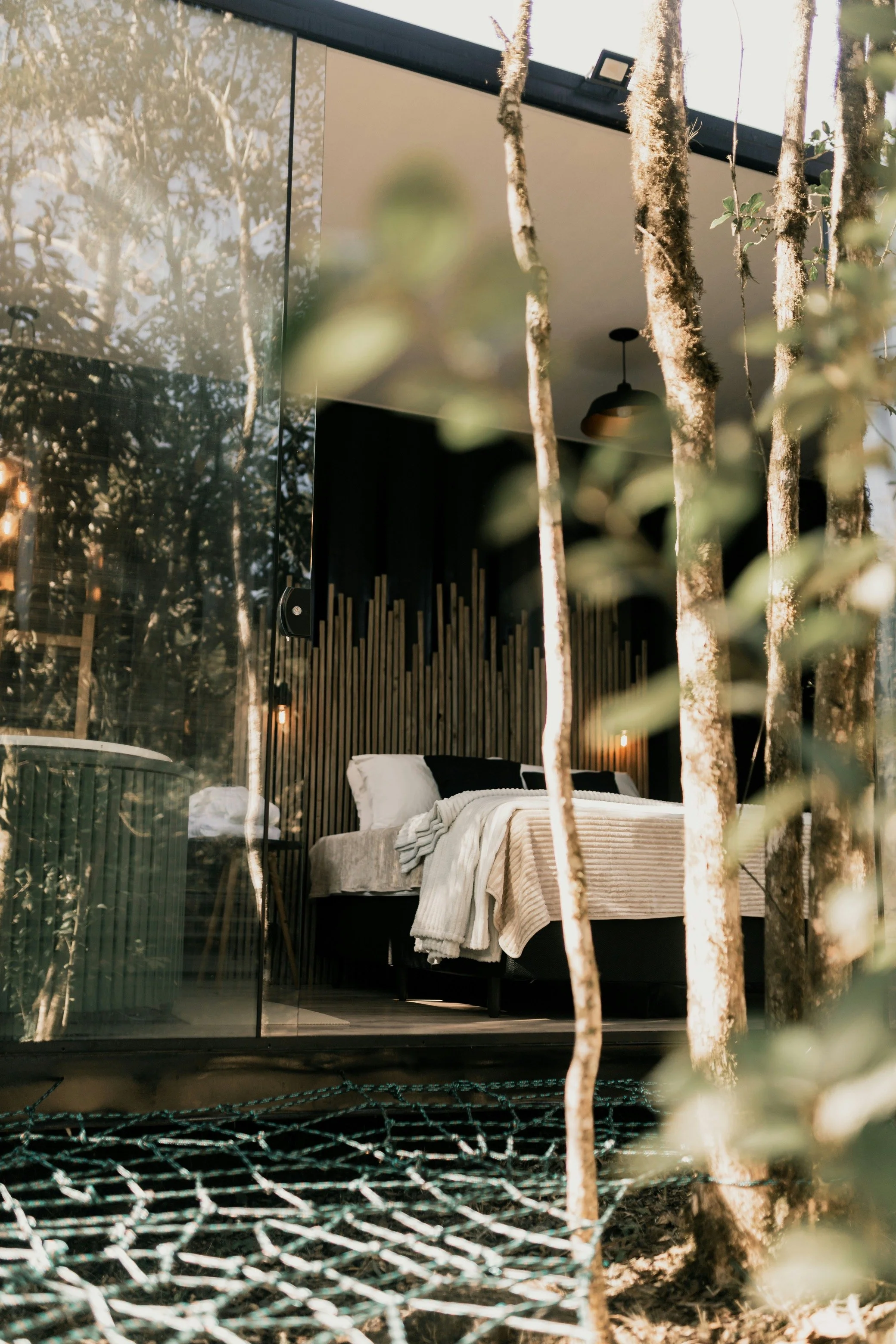 Interior of a bedroom with a bed, blankets, and pillows seen through a glass wall, with hanging branches and leaves in the foreground.