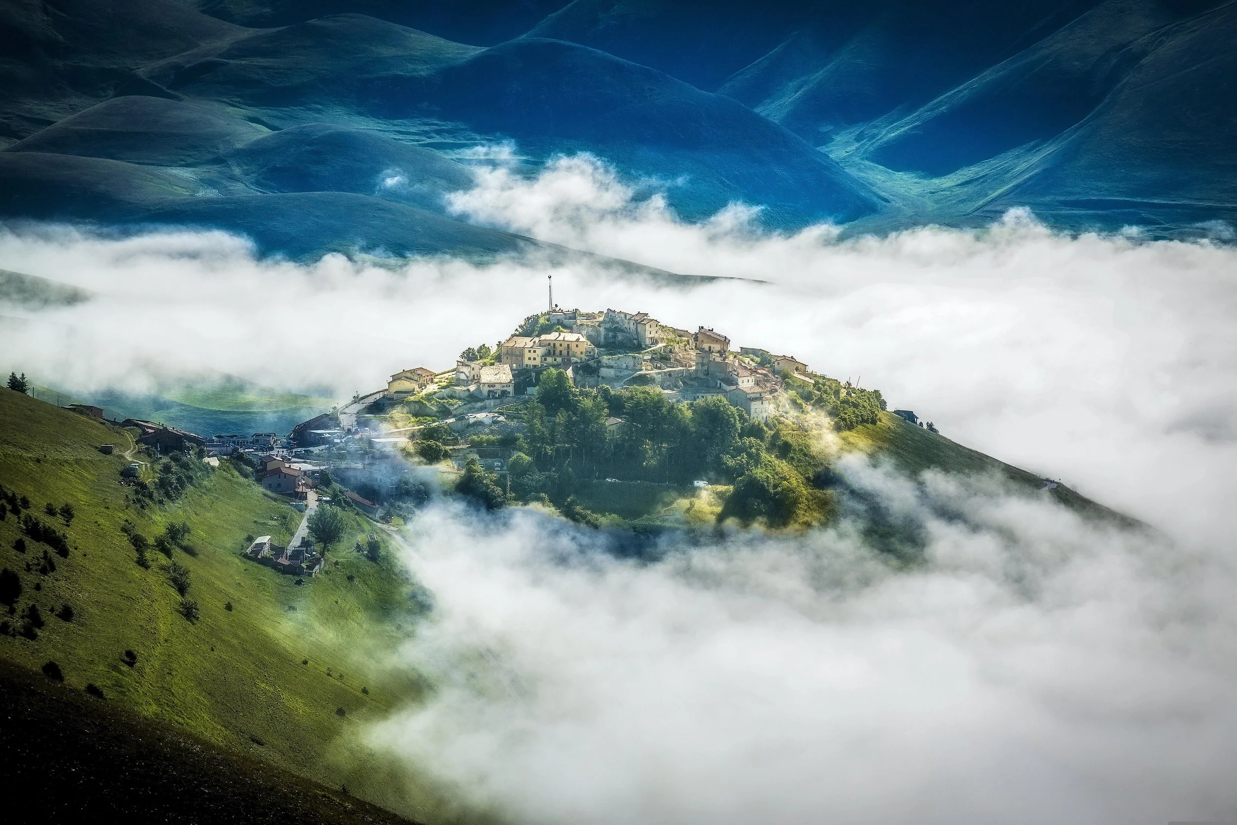 A mountainous landscape with green hills, a small village on a hillside, and clouds filling the valley below.