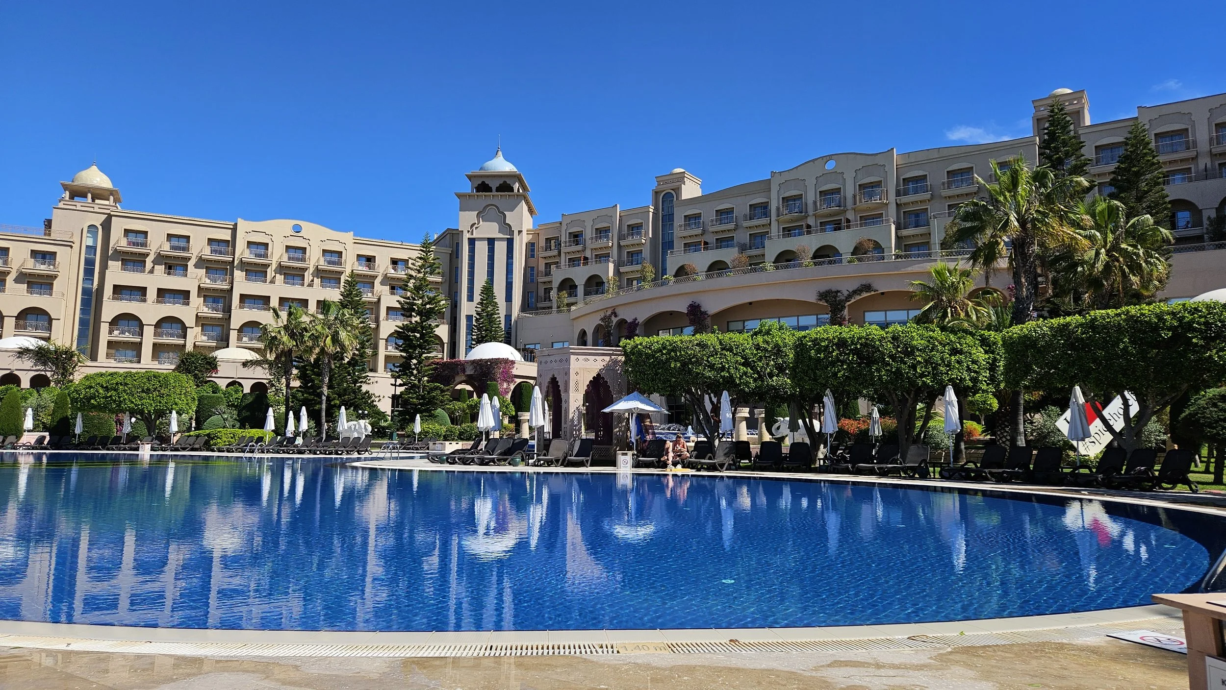 Luxury hotel with multiple floors, water pool with lounge chairs and umbrellas in the foreground, lush greenery, and a clear blue sky.