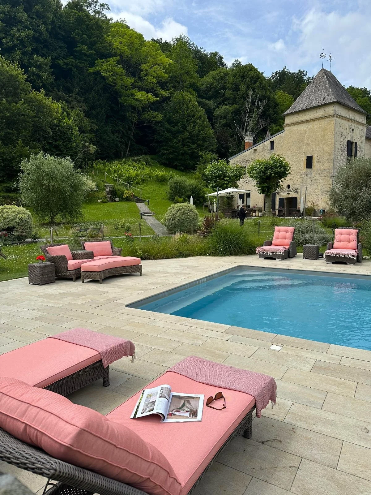 A backyard with a swimming pool and pink cushioned outdoor furniture, situated on a patio with beige tiles. In the background, there is a lush green garden leading up to a stone house with a tower and a garden path, surrounded by trees, under a partly cloudy sky.