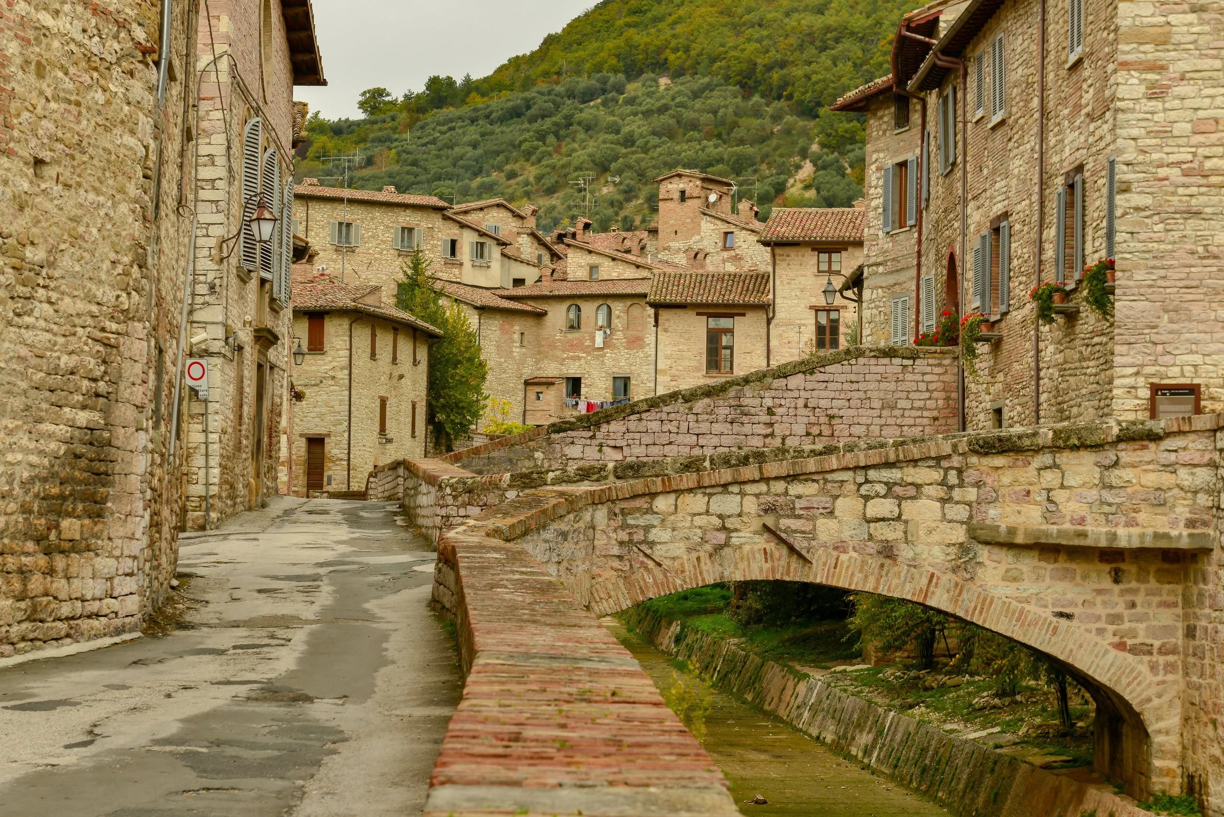 A rustic Italian village with stone buildings, narrow alleyway, arched stone bridge, and lush green hillside in the background.