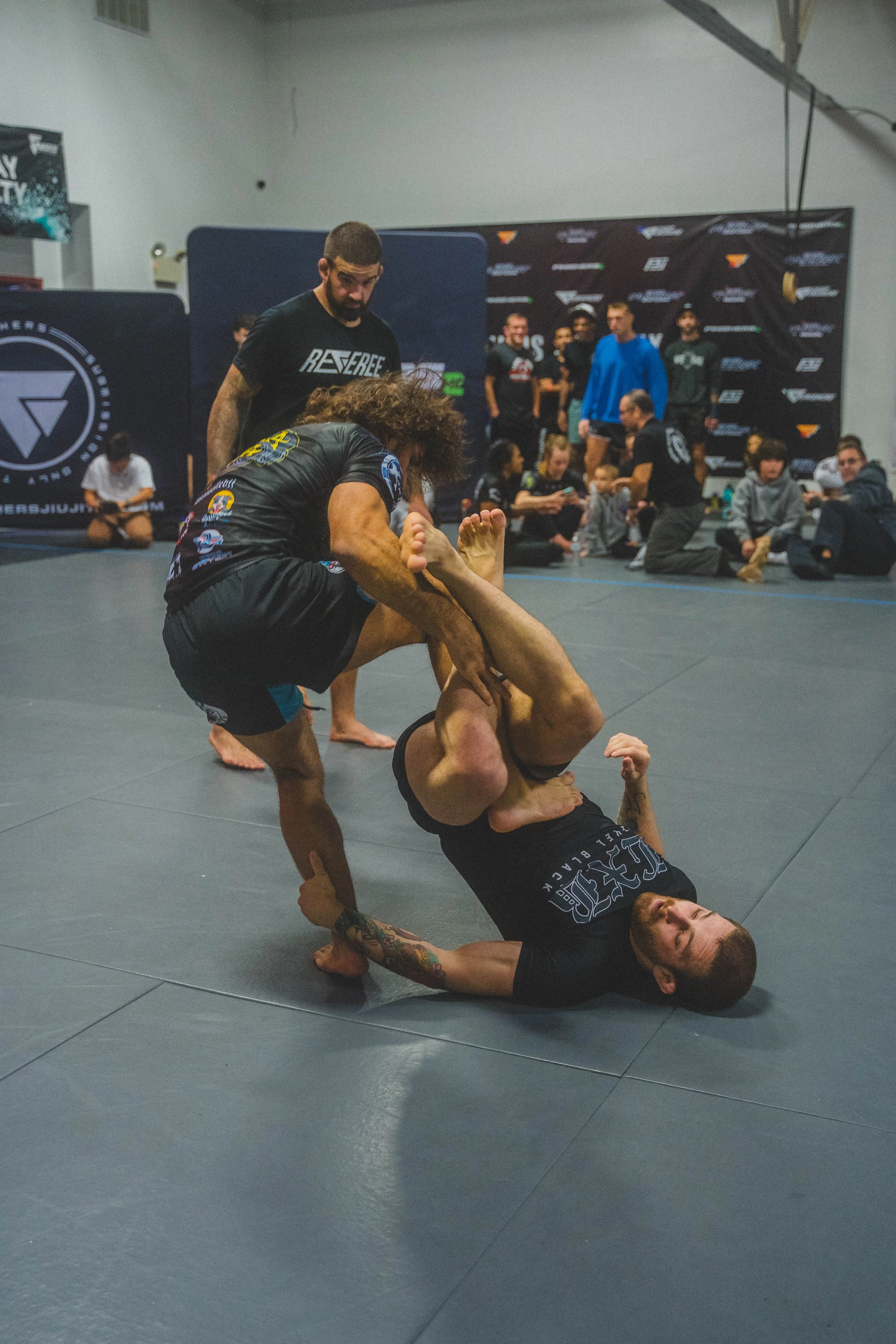 Two men practicing Brazilian Jiu-Jitsu on a gray mat, with one man on his back and the other attempting an arm lock, while a group of people watches in the background.