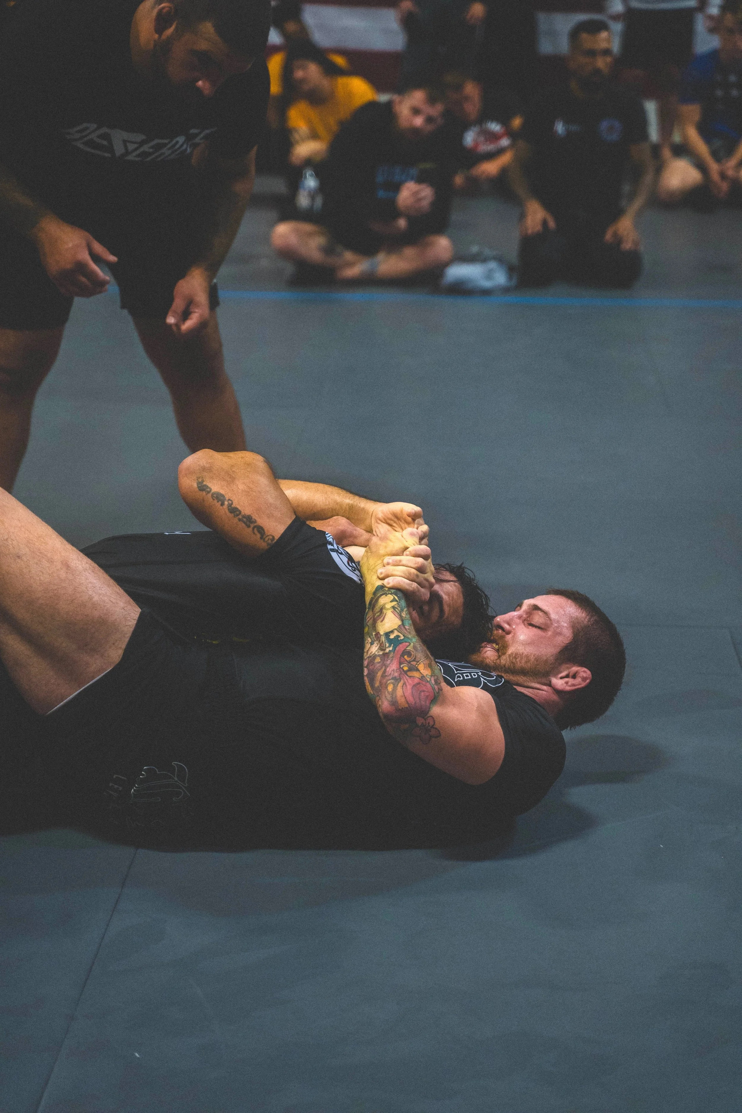 Two men are practicing Brazilian jiu-jitsu on a black mat, one man with tattoos on his arms is on his back attempting a grappling move while the other man is above him in a dominant position. In the background, a group of people are sitting on the ma