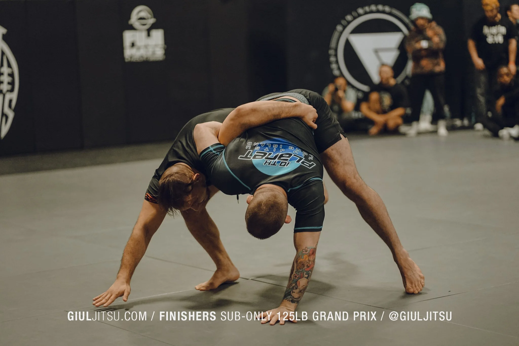 Two men practicing Brazilian Jiu-Jitsu on a mat, with one man gripping the other's arm and neck in a grappling position. Several people watch in the background, some sitting and others standing, in a martial arts gym with black walls and logos.