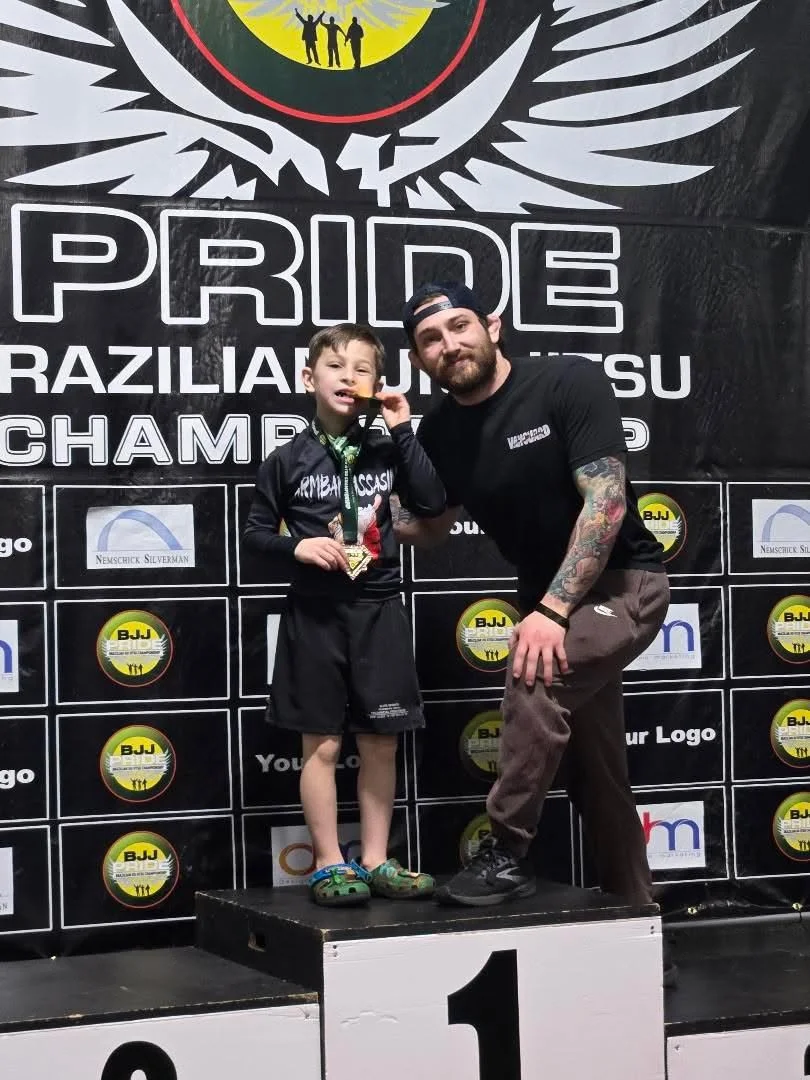 A young boy and a man are standing on a winners' podium after a Brazilian Jiu-Jitsu competition. The boy is holding a gold medal and a small trophy, smiling at the camera. They are in front of a large banner with the words "PRIDE" and "Brazilian Jiu-Jitsu Championship," along with various sponsor logos. The boy is wearing a black Jiu-Jitsu gi, and the man is dressed in casual athletic clothing.