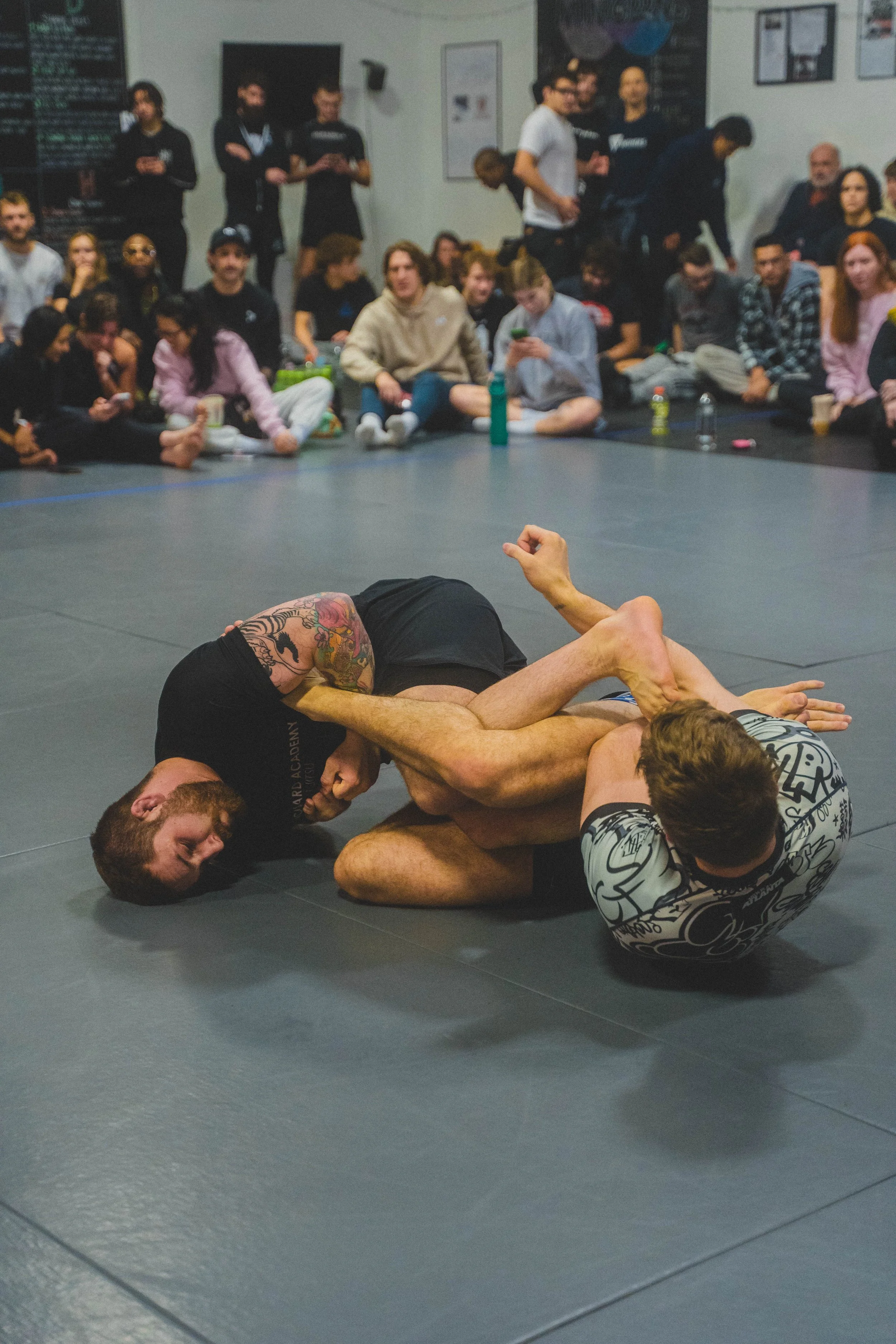 Two men engage in Brazilian jiu-jitsu on a gray mat, with a diverse group of spectators sitting around and watching.
